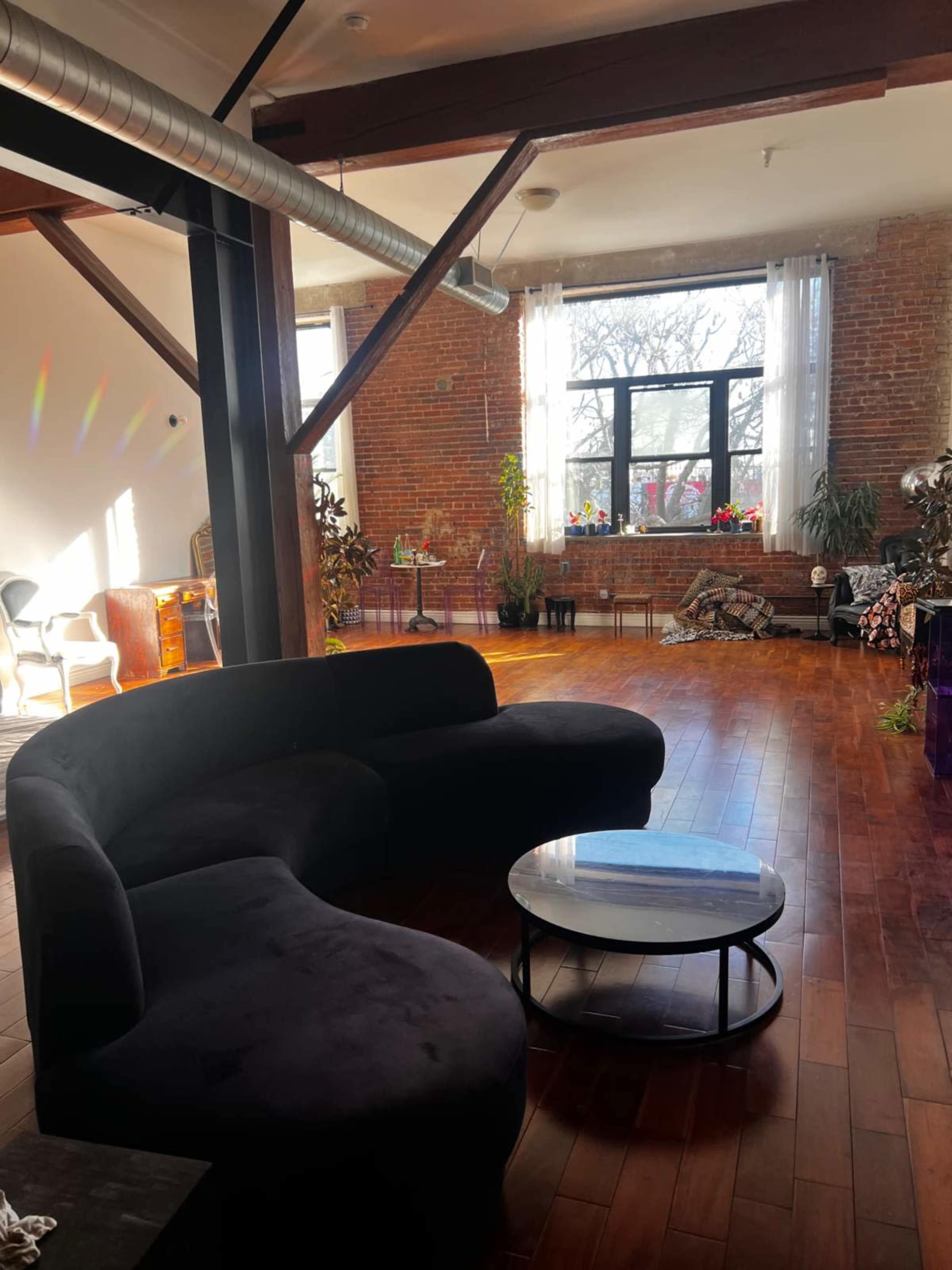 The image shows a spacious interior with exposed brick walls, large windows allowing natural light, and a curved black couch facing a round coffee table on a wooden floor.