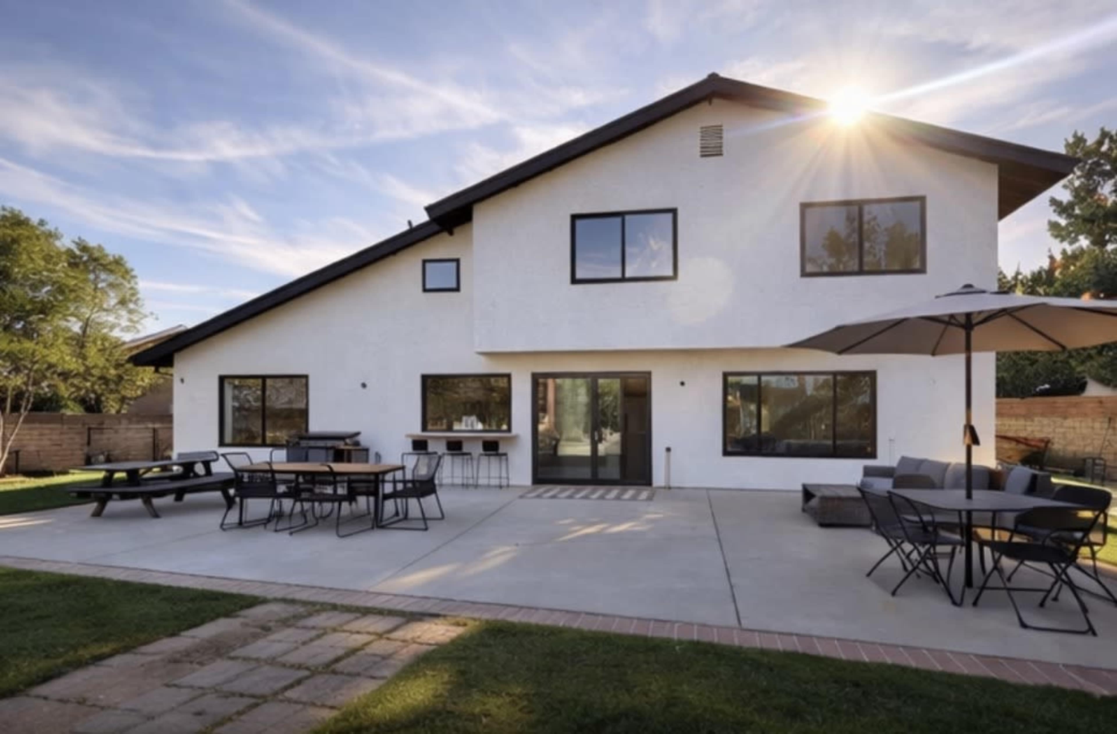 The image shows a modern white house with large windows, featuring an outdoor patio area with tables, chairs, and an umbrella in a landscaped yard.