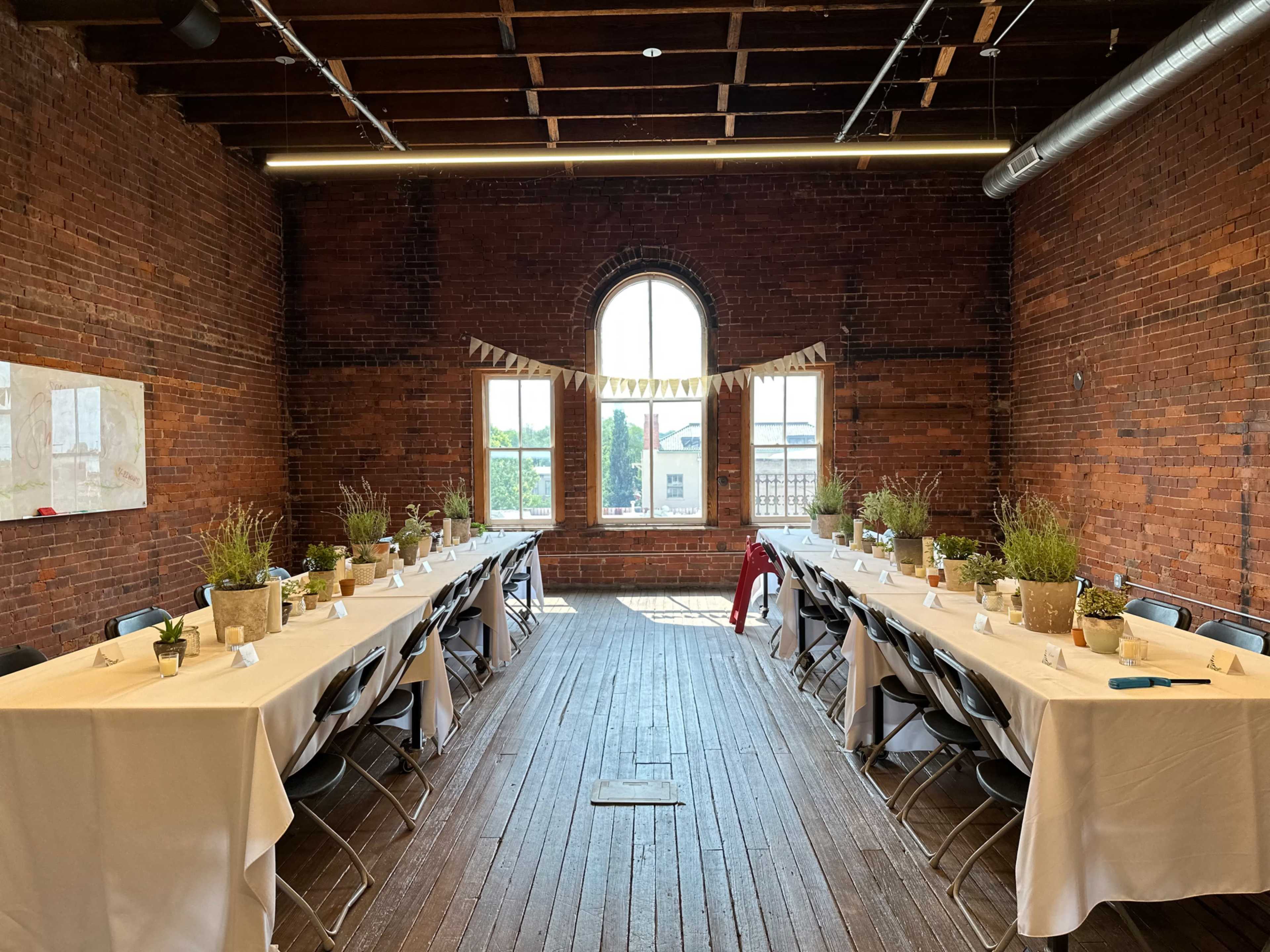 The image shows a rustic brick room set up for a meal with two long tables covered in white tablecloths, each adorned with potted plants and centerpieces, and large windows allowing natural light to enter.