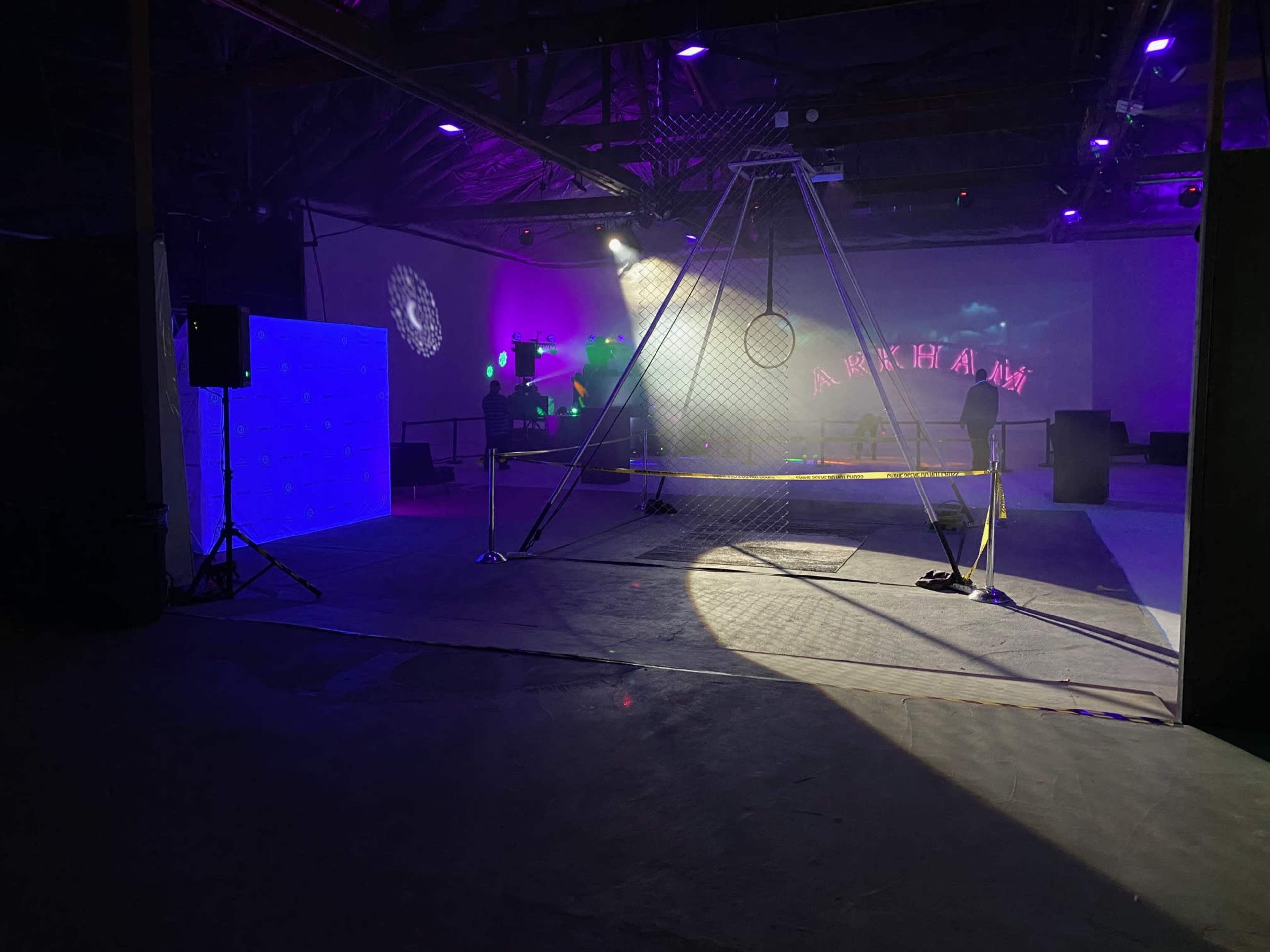 The image shows an industrial space set up for an event, featuring a large mesh structure with a hoop, a blue illuminated backdrop, and various lights in the background.