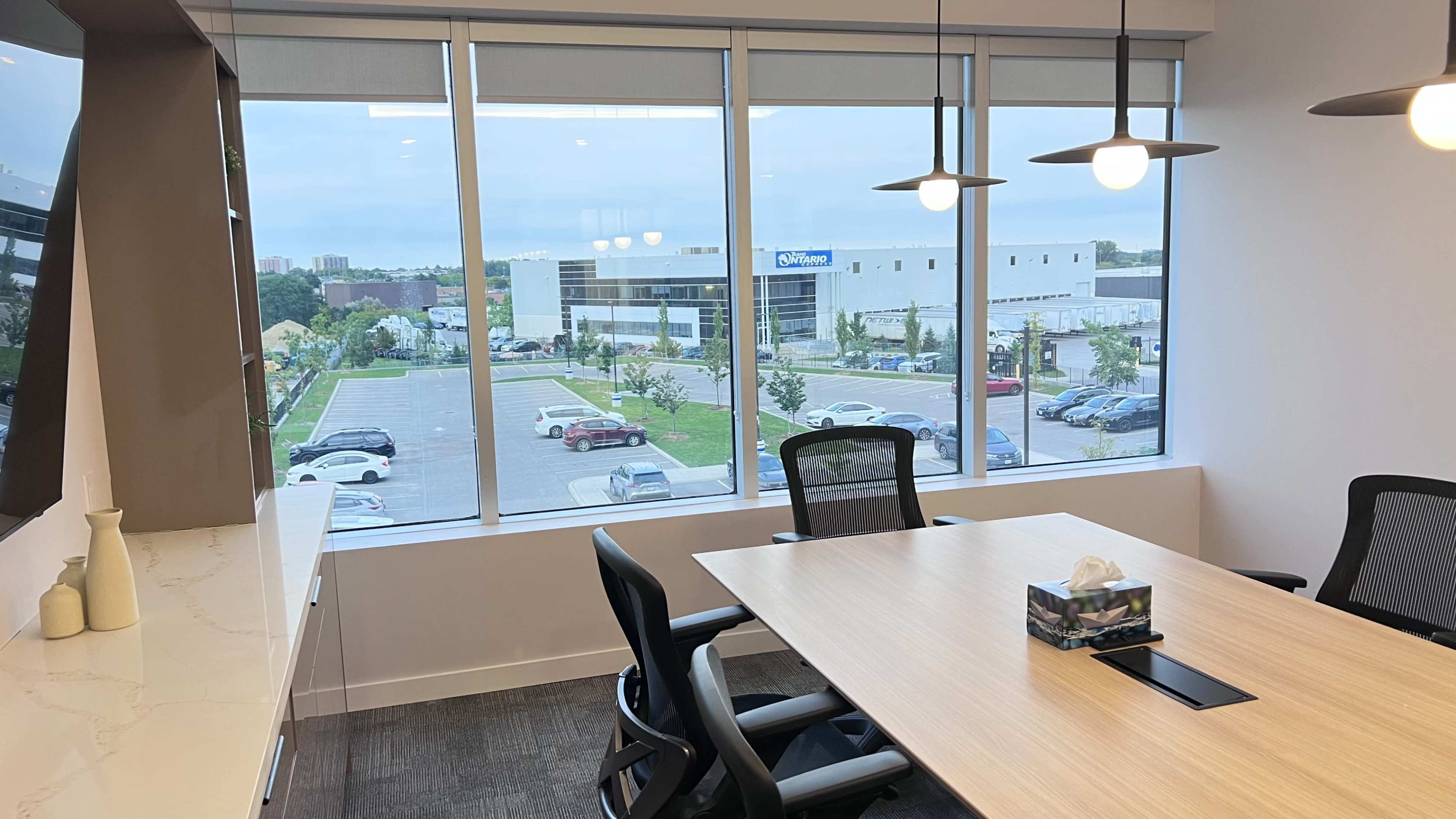 A modern conference room features a large wooden table and chairs, with a view of a parking lot and commercial buildings through large windows.