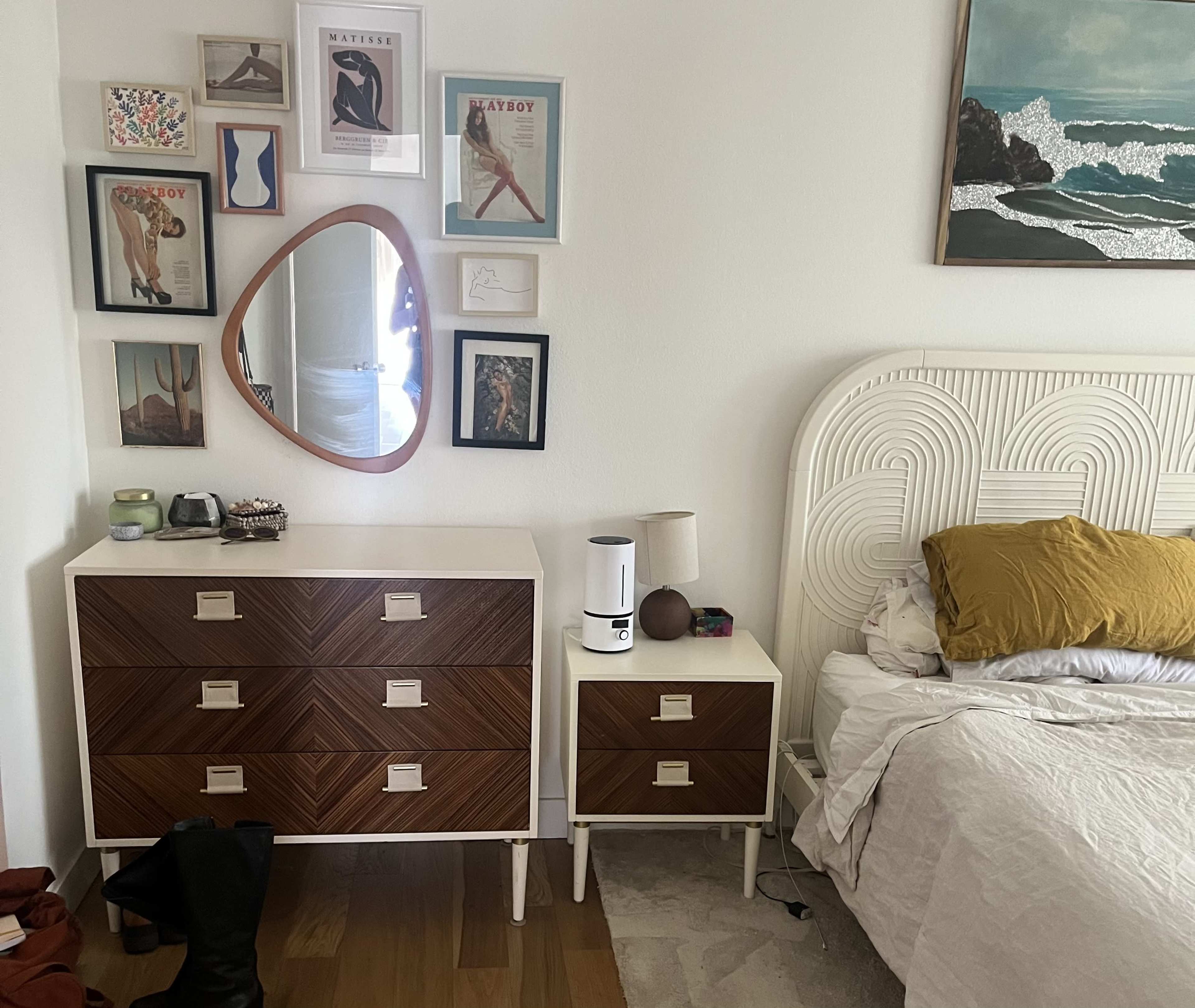 A bedroom with a white bed, a wooden dresser with four drawers, a bedside table, a mirror, and several framed artworks on the wall.