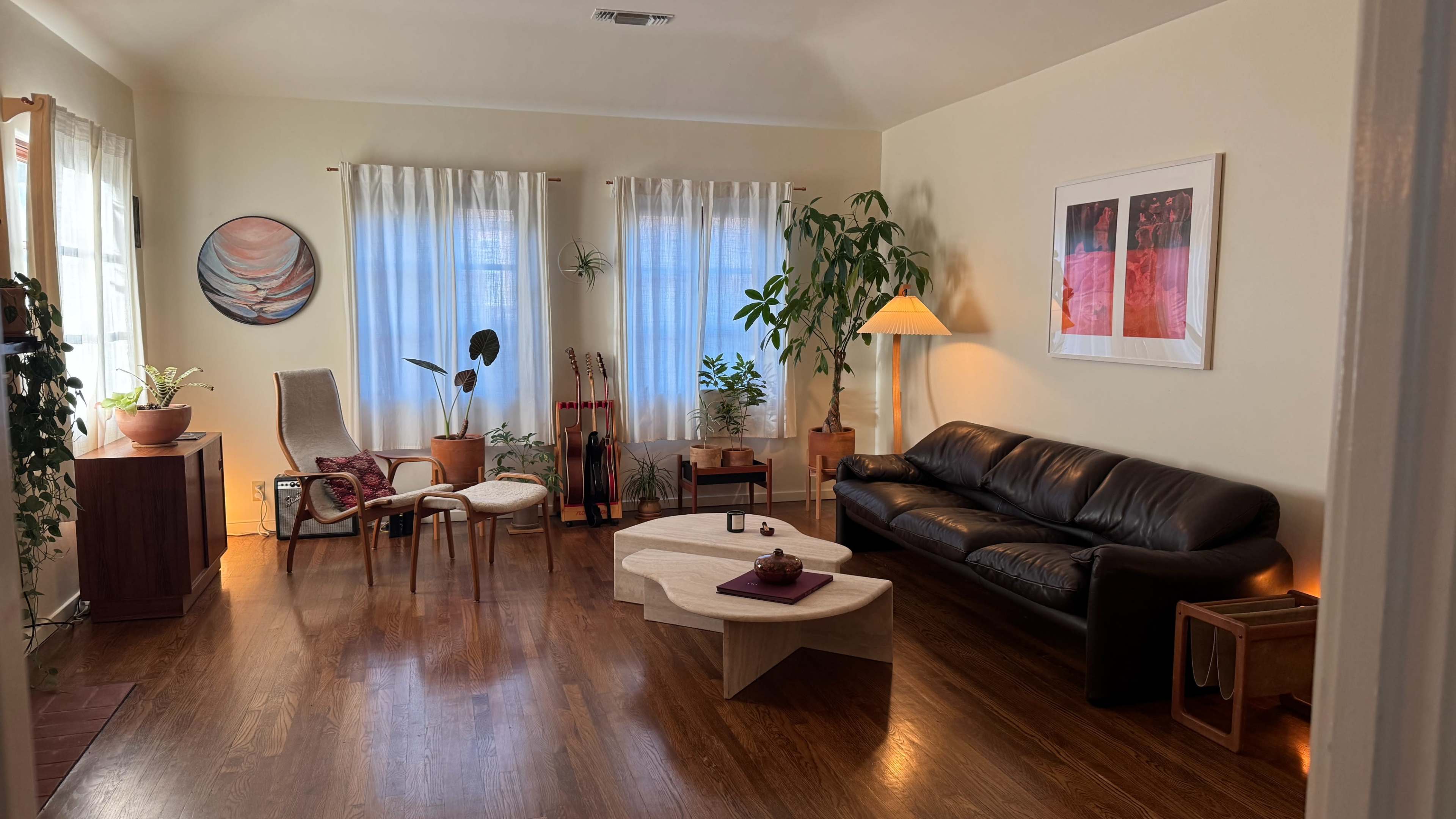 The room features a seating area with a black leather sofa, wooden coffee table, and various plants, illuminated by natural light from large windows.
