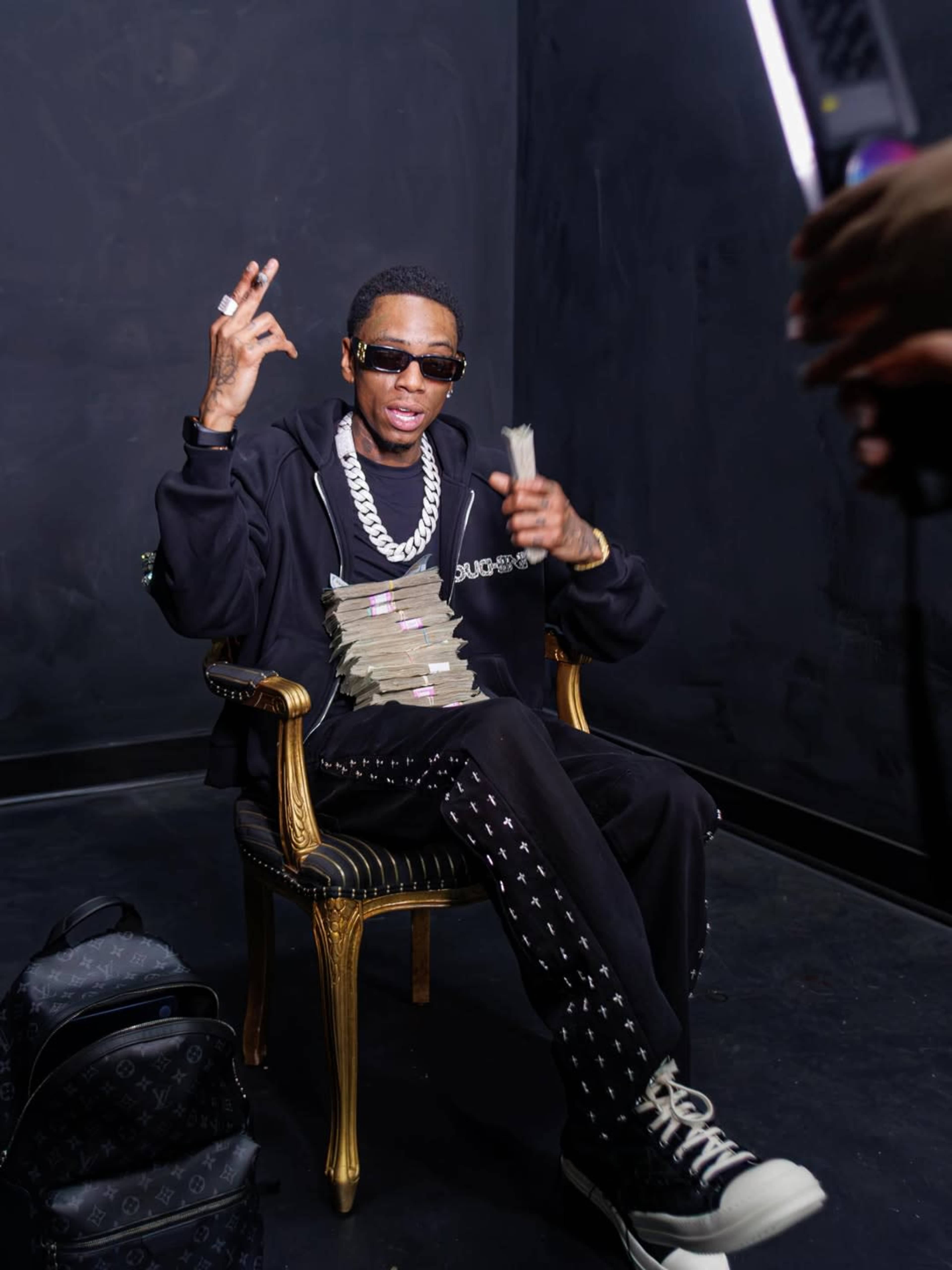 A man wearing sunglasses and a black outfit sits in a chair, holding stacks of cash while making a hand gesture.