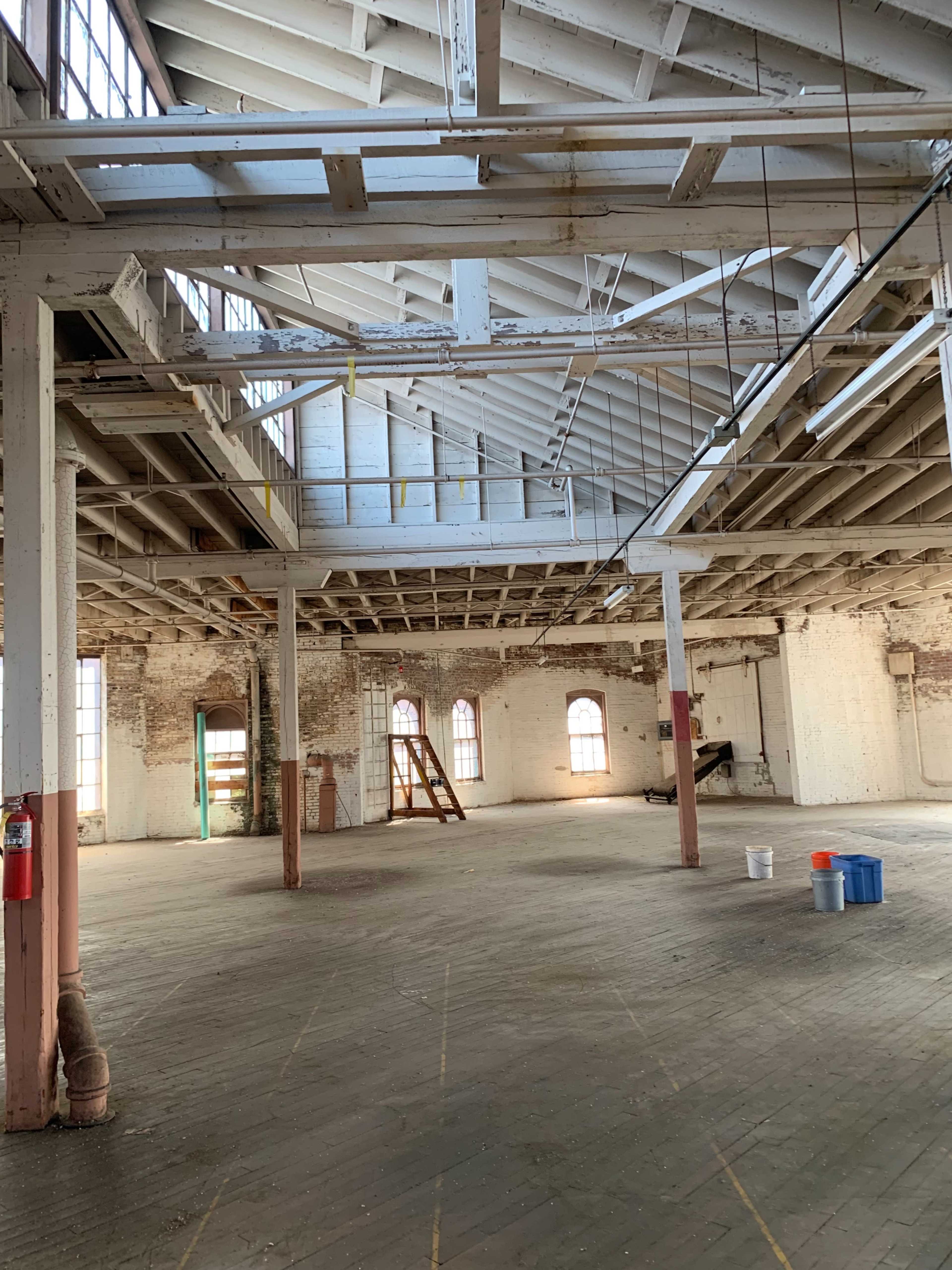 The image shows an empty, open space in a warehouse with exposed wooden beams and large windows.