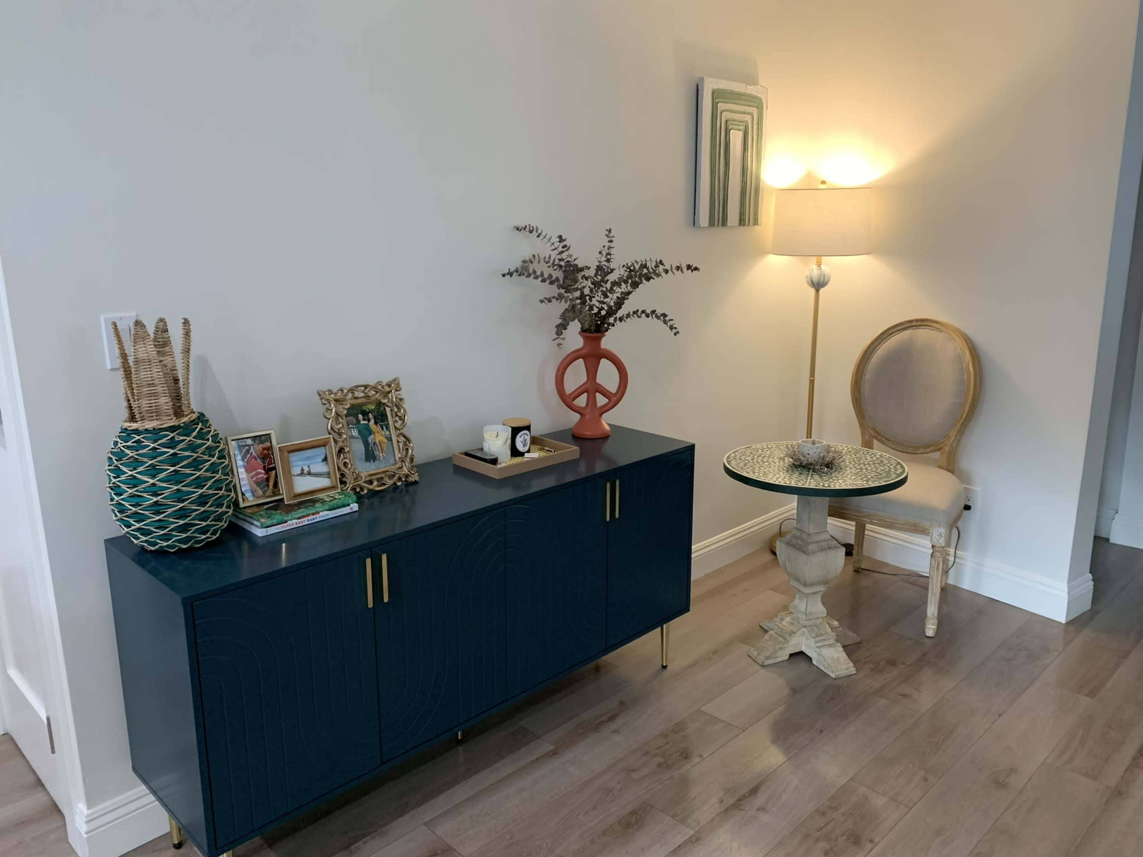 The image shows a modern living space featuring a dark blue sideboard with decorative items, a peace sign sculpture, and a side table with a patterned top beside a lamp on a light-colored wall.