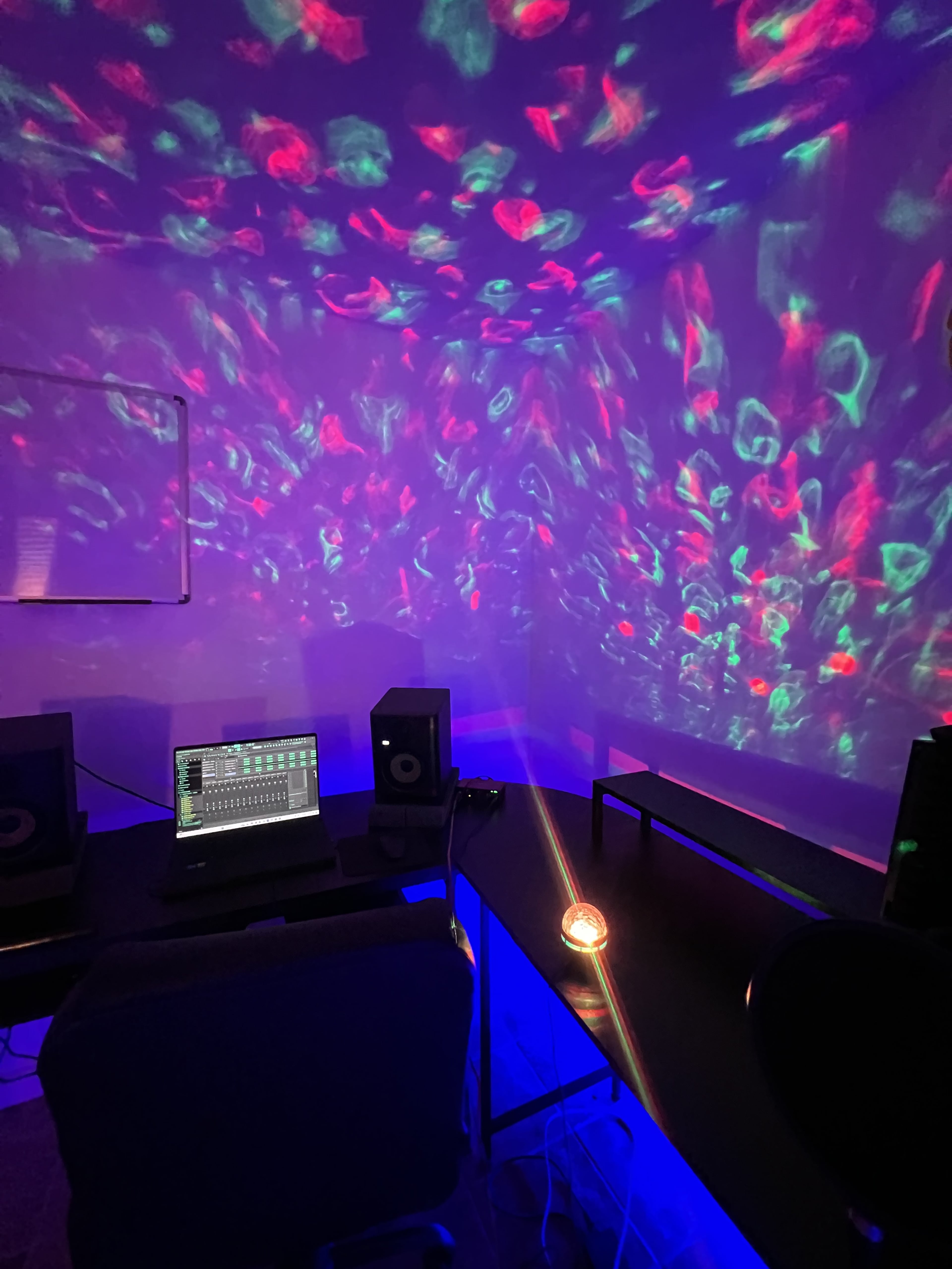 A dimly lit music studio with a computer, speakers, and colorful projections on the walls.