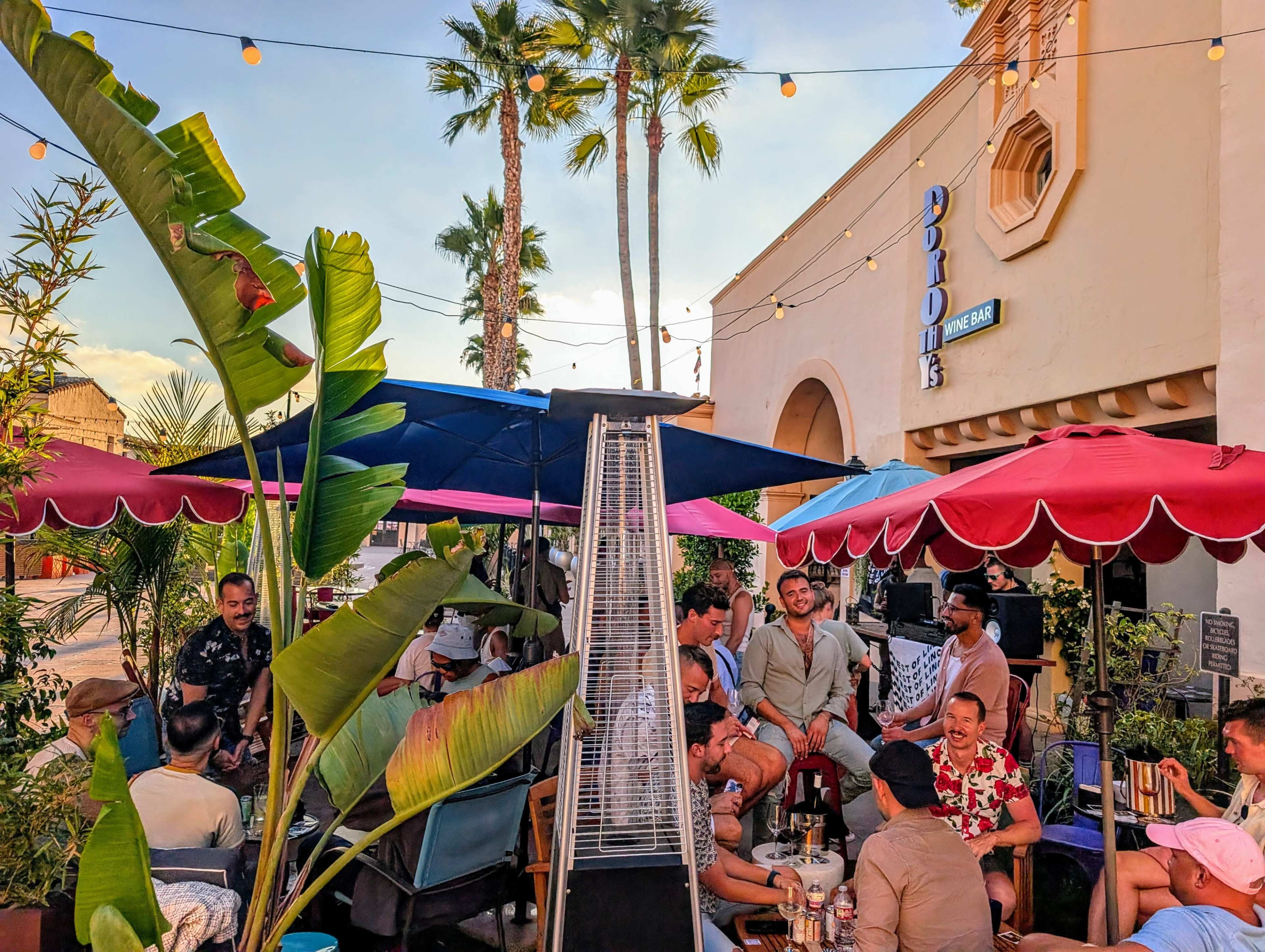 A lively outdoor bar scene features patrons gathered under colorful umbrellas, surrounded by palm trees and string lights, enjoying food and drinks.