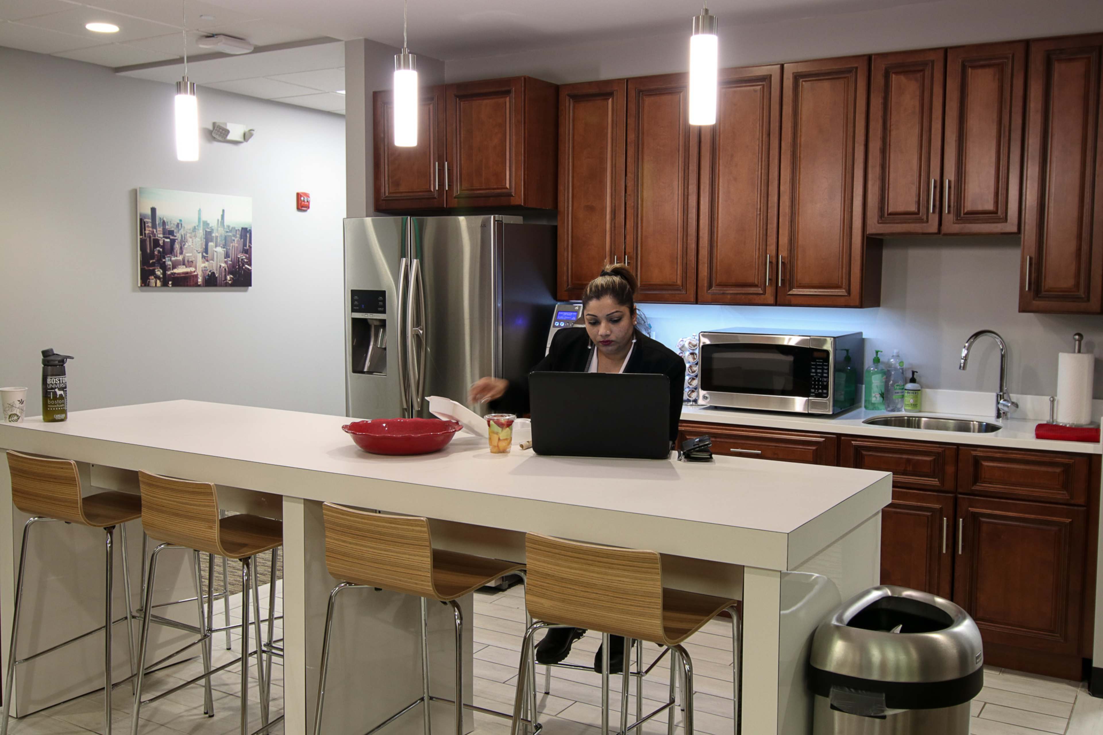 A woman works on a laptop at a kitchen island in a modern kitchen with wooden cabinets and stainless steel appliances.