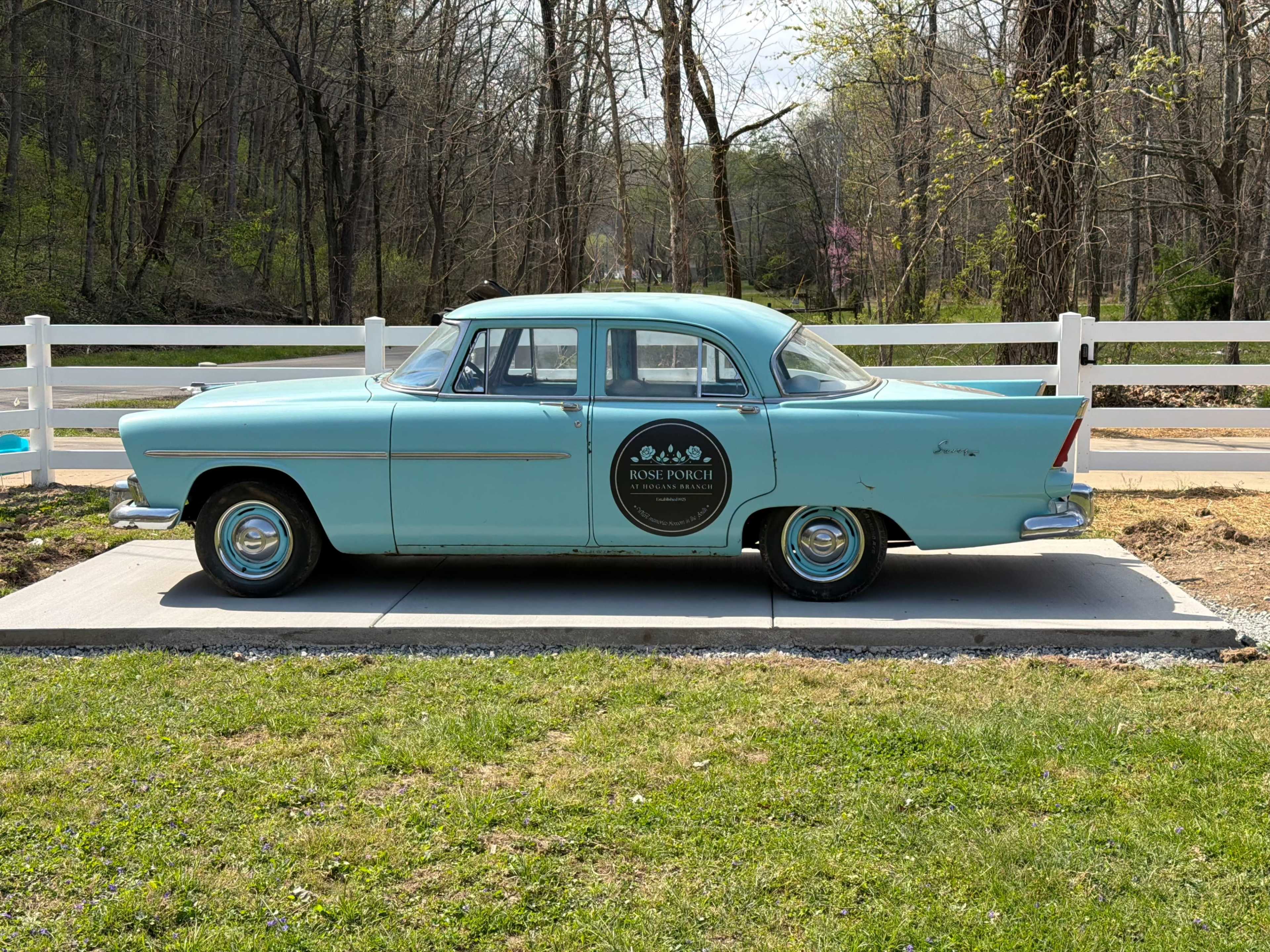A vintage turquoise car is parked on a concrete slab beside a white wooden fence in a wooded area.