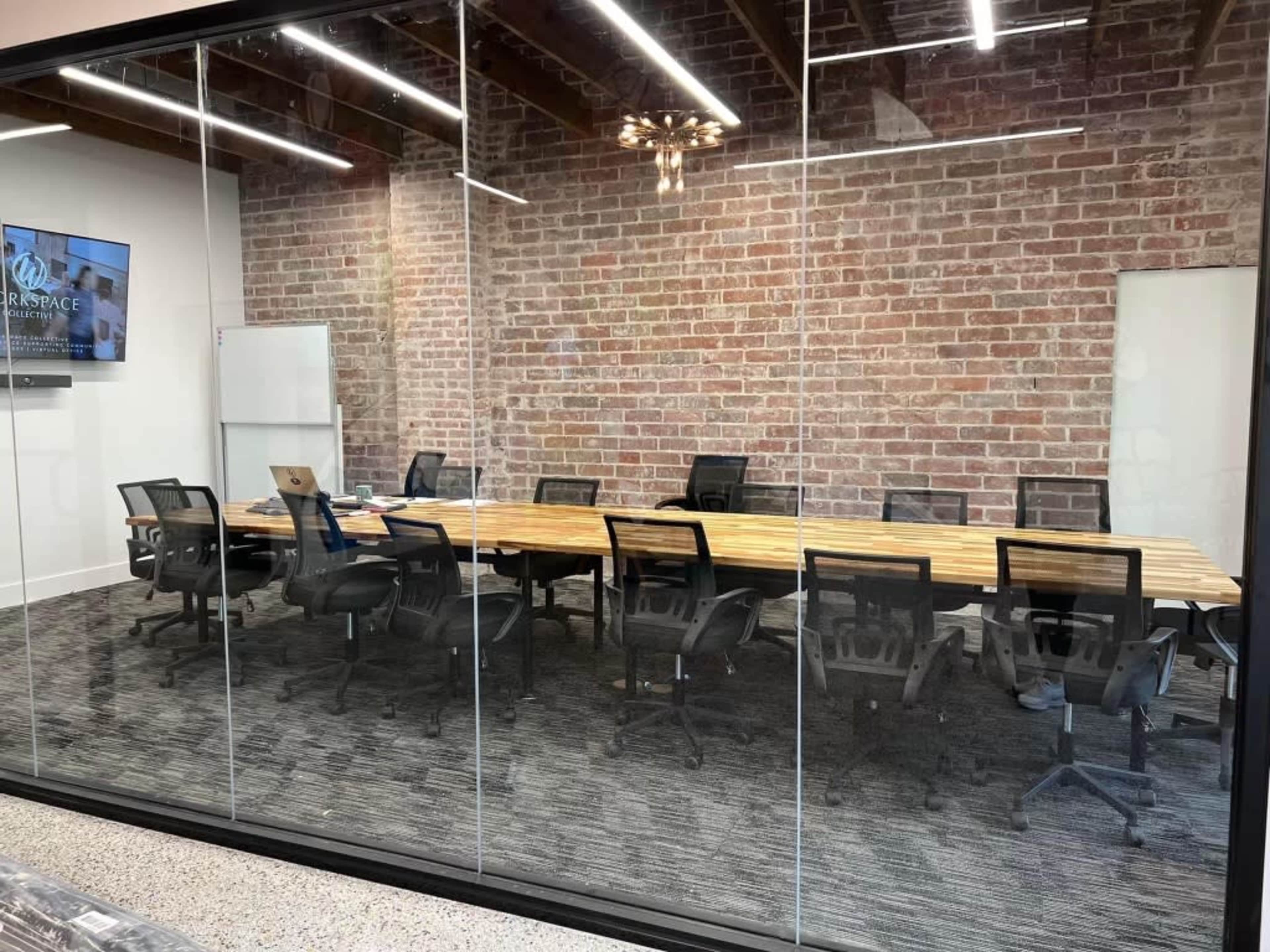 The image shows a modern conference room with a large wooden table surrounded by black office chairs, all enclosed by glass walls and featuring exposed brick and contemporary lighting.