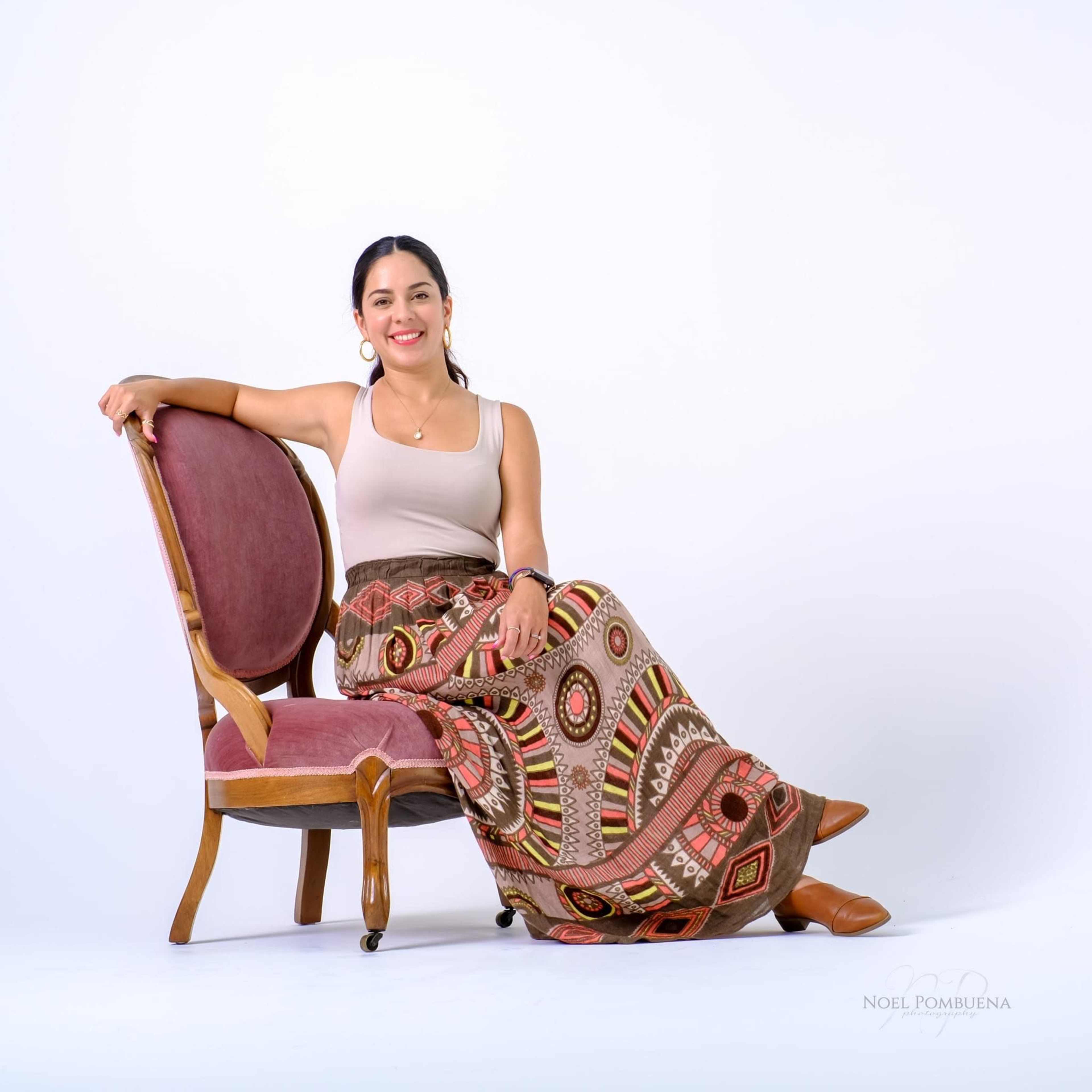 A woman wearing a beige tank top and a patterned skirt sits on a vintage chair against a plain white background.