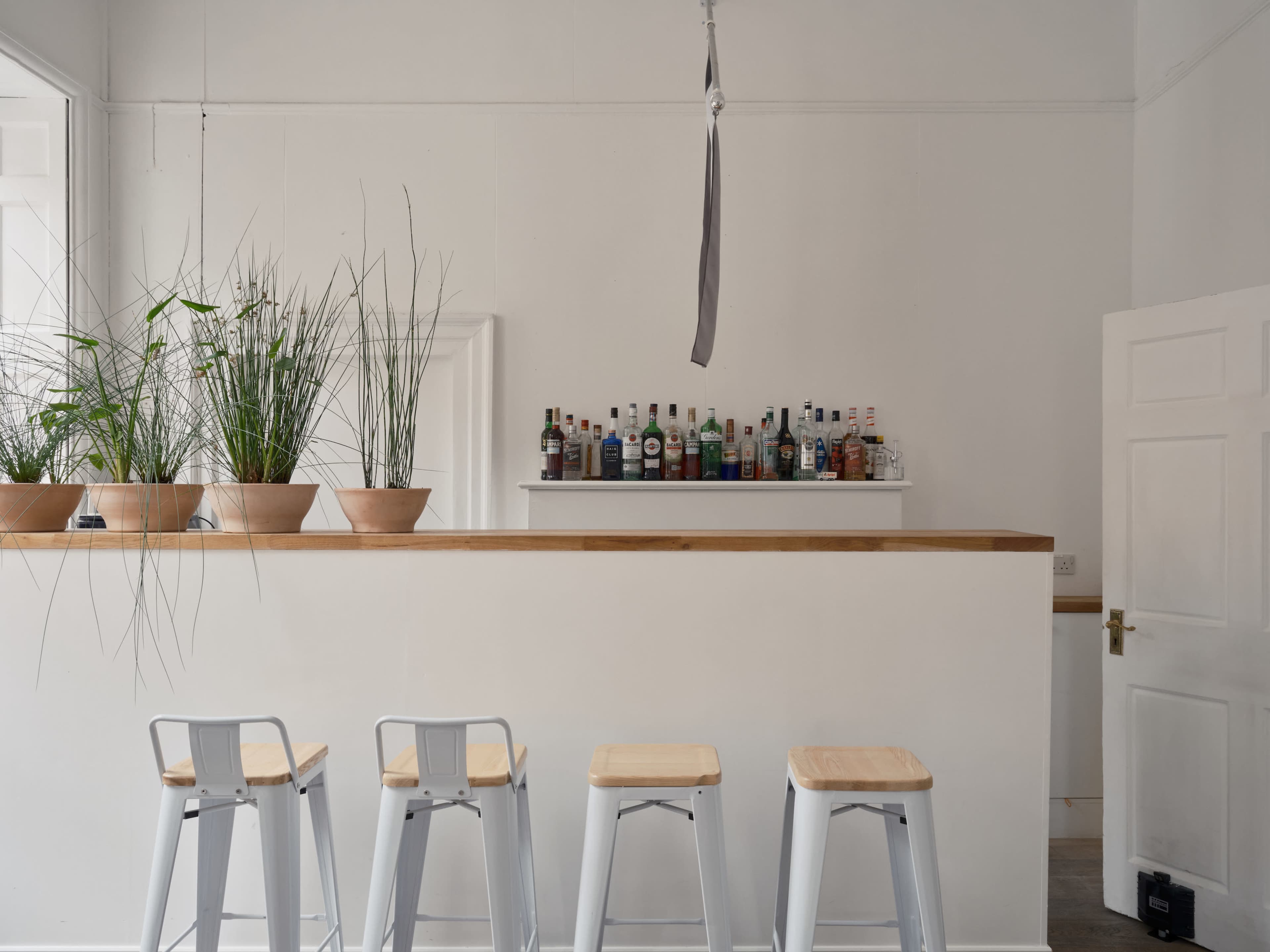A minimalist bar area features a simple counter with potted plants and a row of bottles displayed on a shelf above, accompanied by four metal stools.