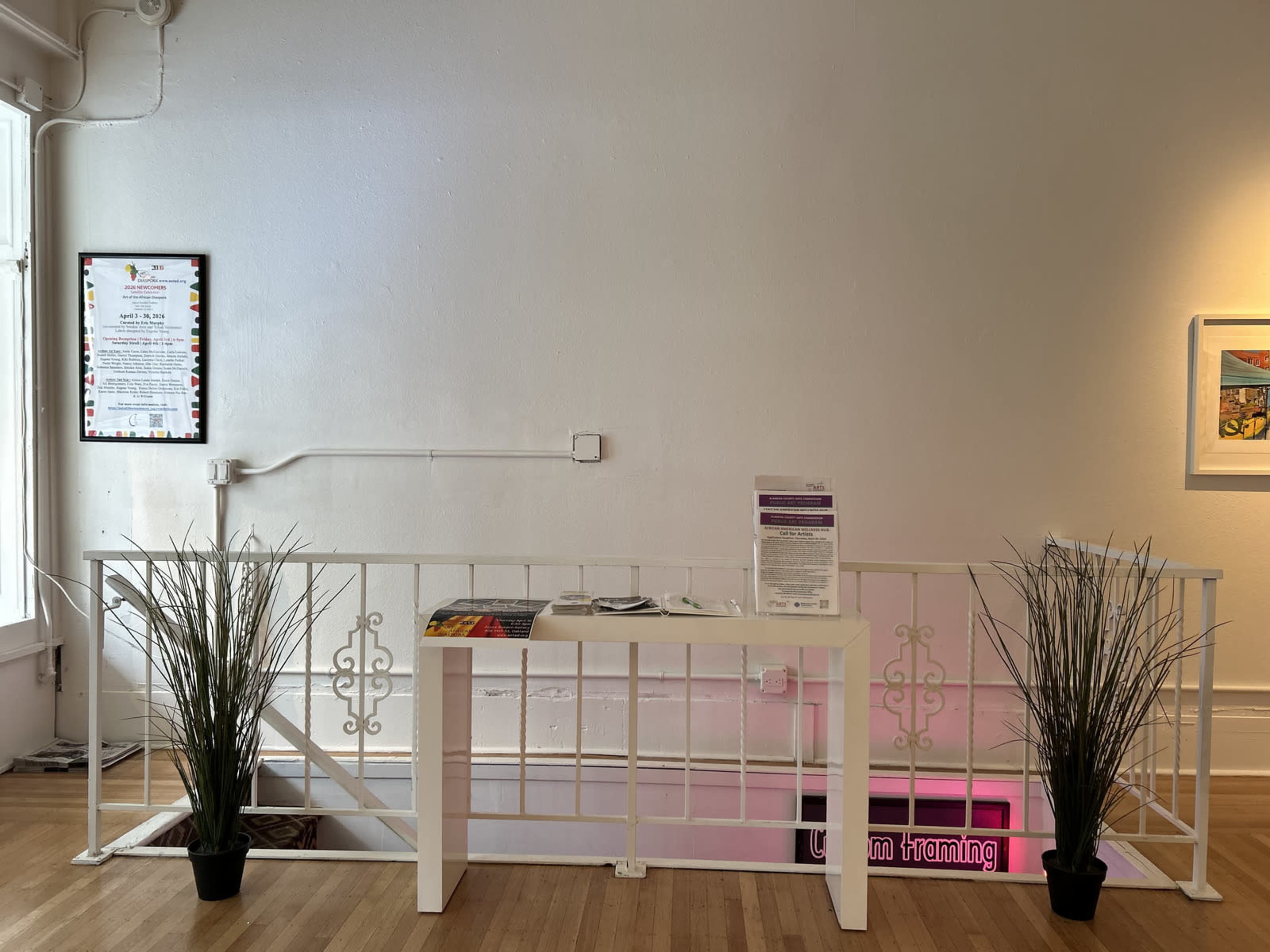 The image shows a simple white railing with a table in the center, flanked by potted plants, in a bright indoor space featuring a menu on the wall.