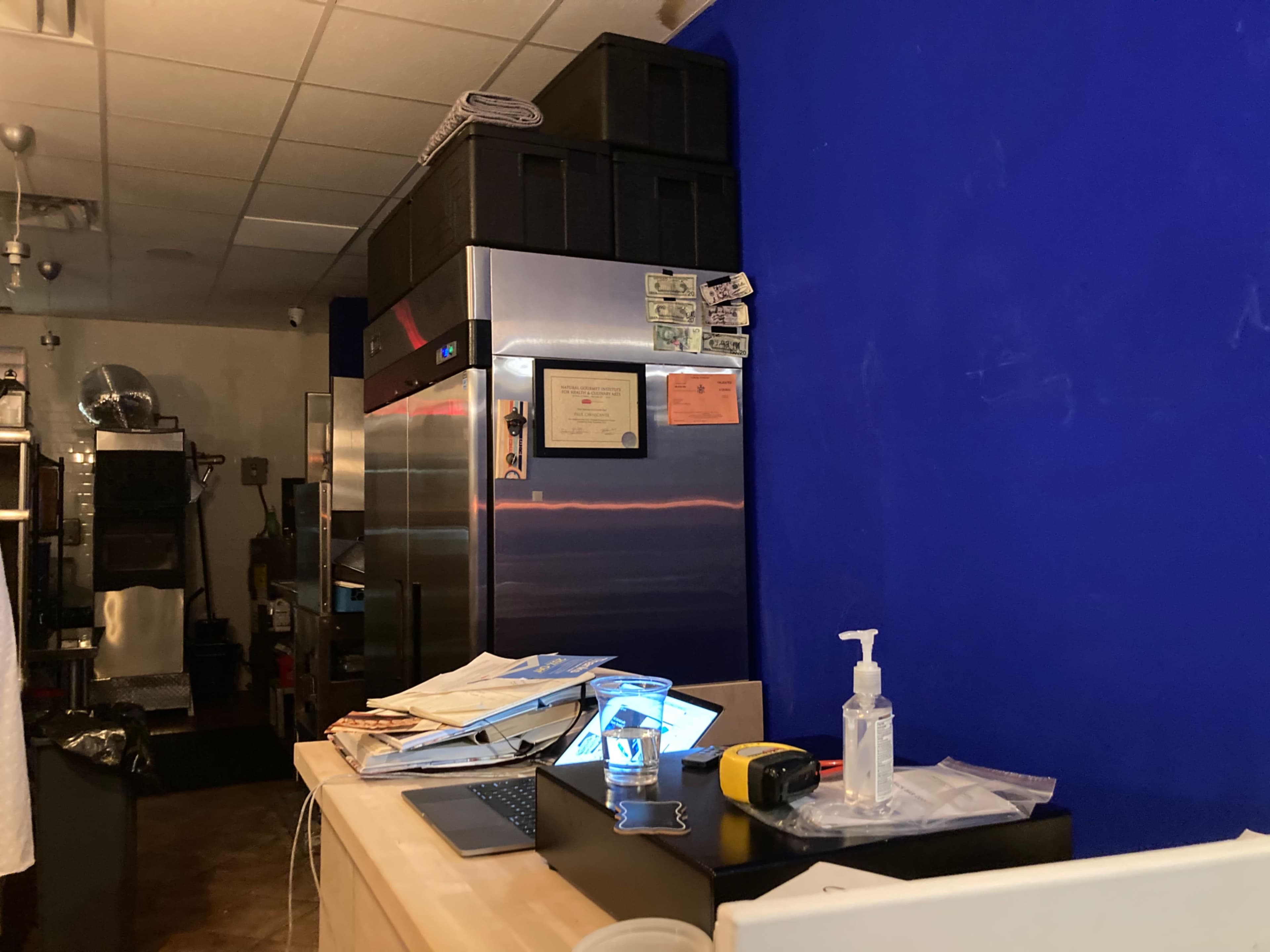 The image shows a kitchen counter cluttered with papers, a laptop, and a sanitizer bottle, with a stainless steel refrigerator in the background against a blue wall.