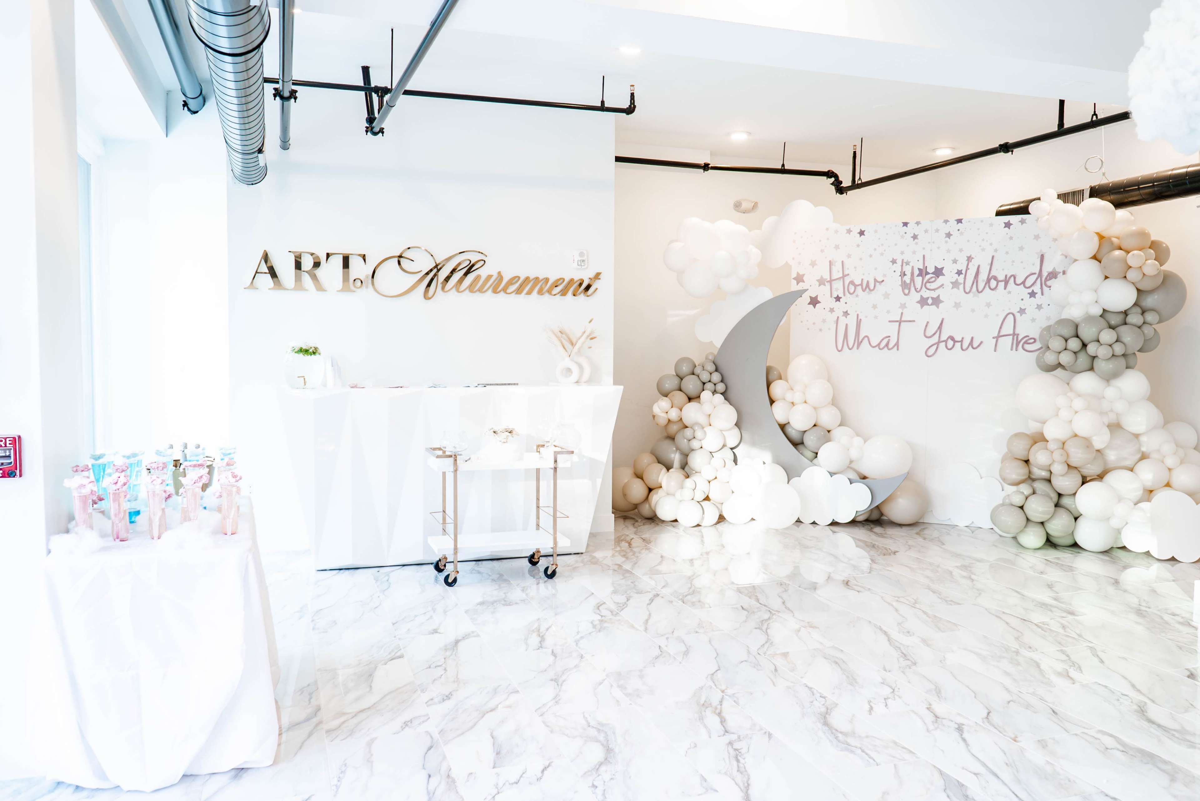 A brightly lit event space with a white marble floor, decorated with a large "ART Allurement" sign, a reception area, and a backdrop of balloons in various colors.