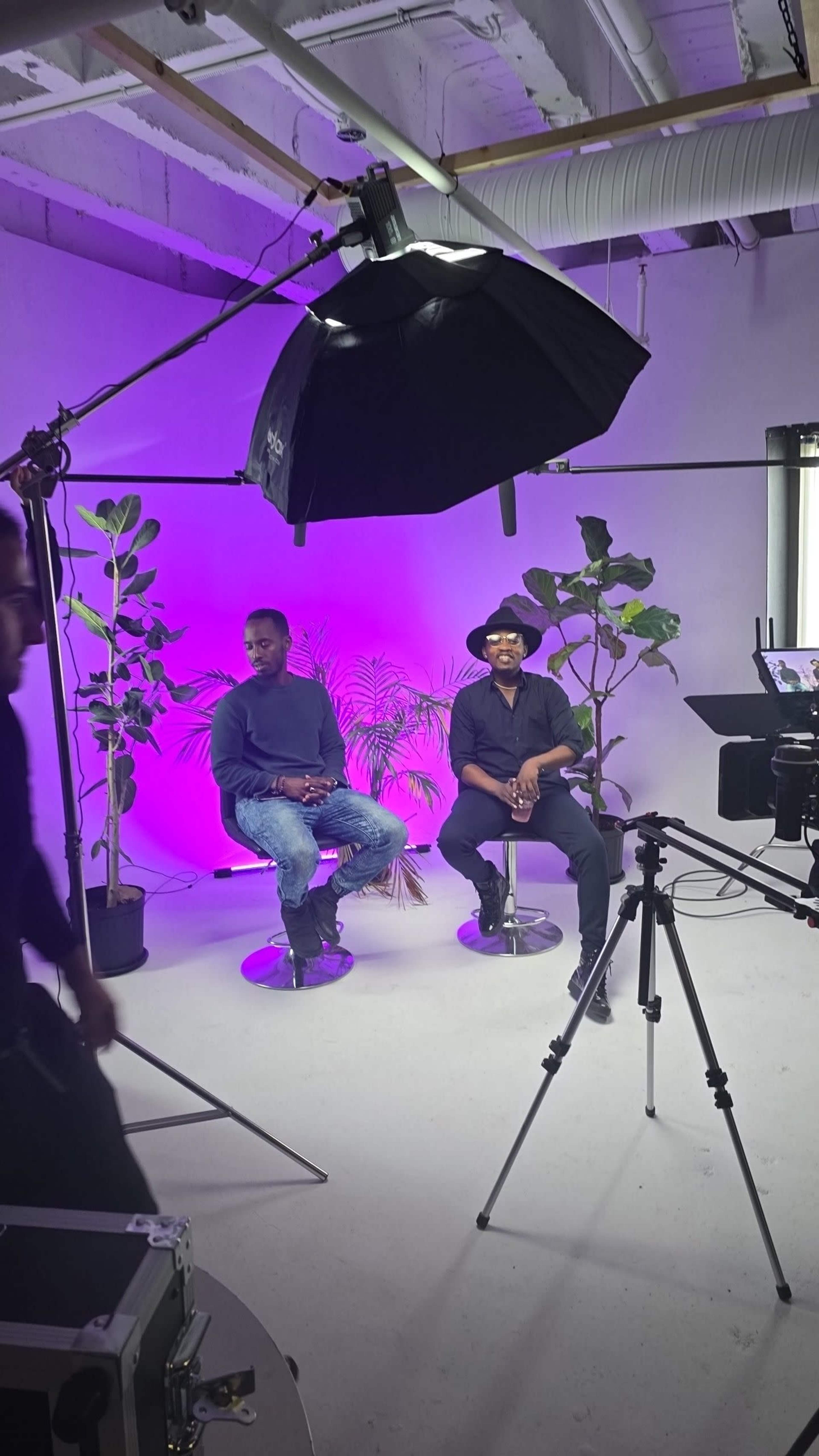 Two men sit on swivel chairs in a studio with purple lighting and potted plants while a camera and lighting equipment are set up nearby.