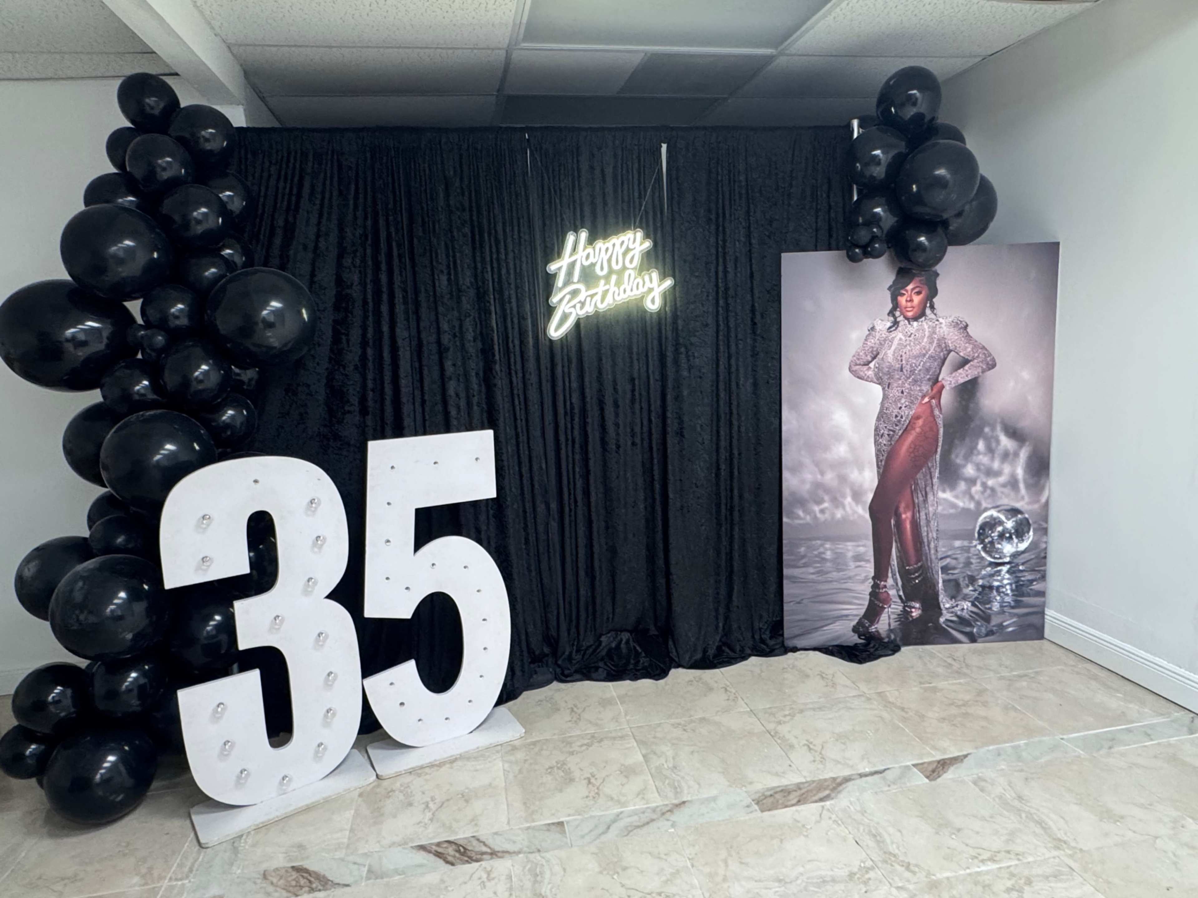 The image shows a birthday celebration setup featuring a backdrop with black curtains, a neon "Happy Birthday" sign, large white illuminated numbers "35," and a photo of a person posing next to a disco ball, surrounded by black balloons.