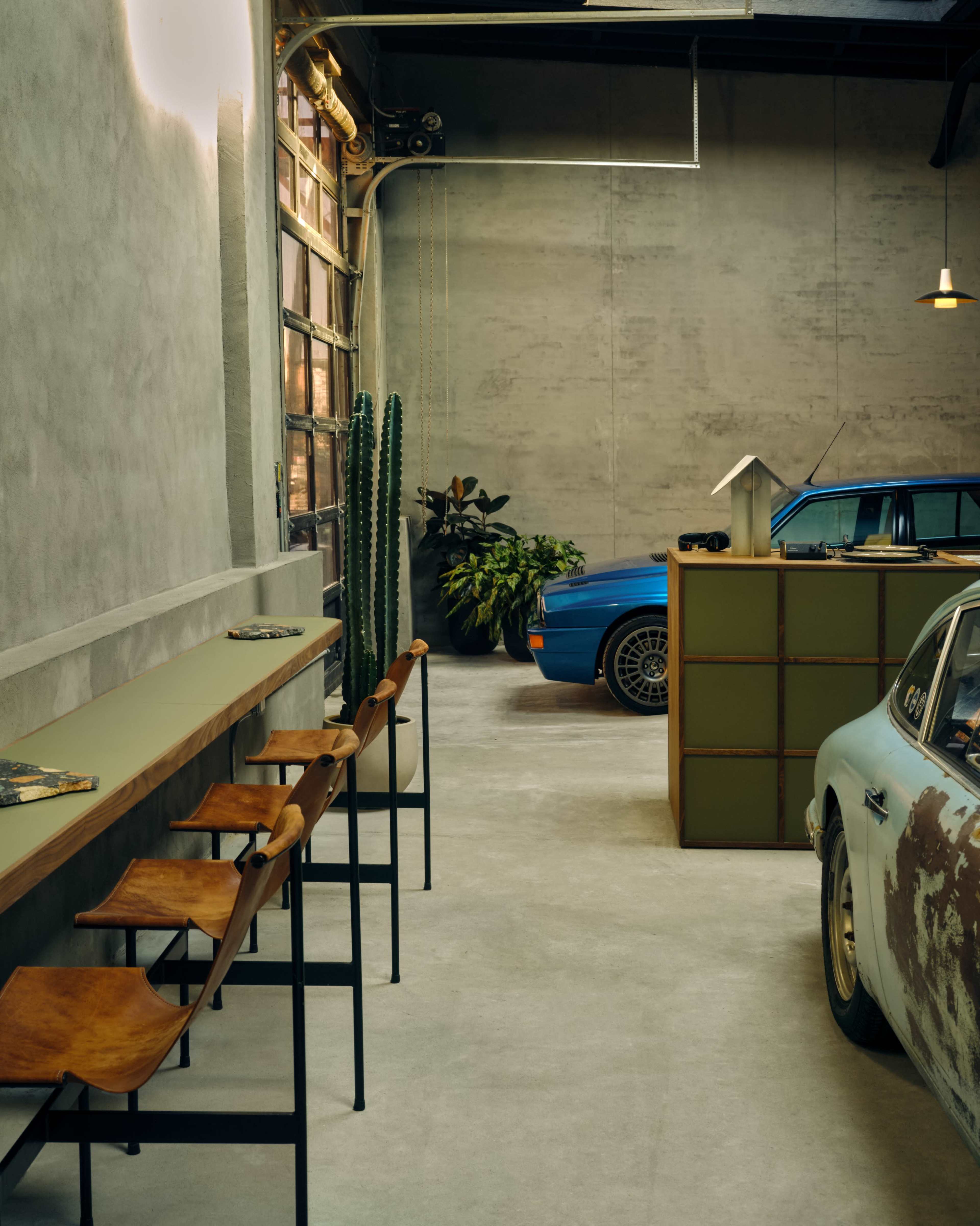 The image depicts an industrial-style garage with two vintage cars, a seating area with metal and leather stools, and potted plants along the walls.