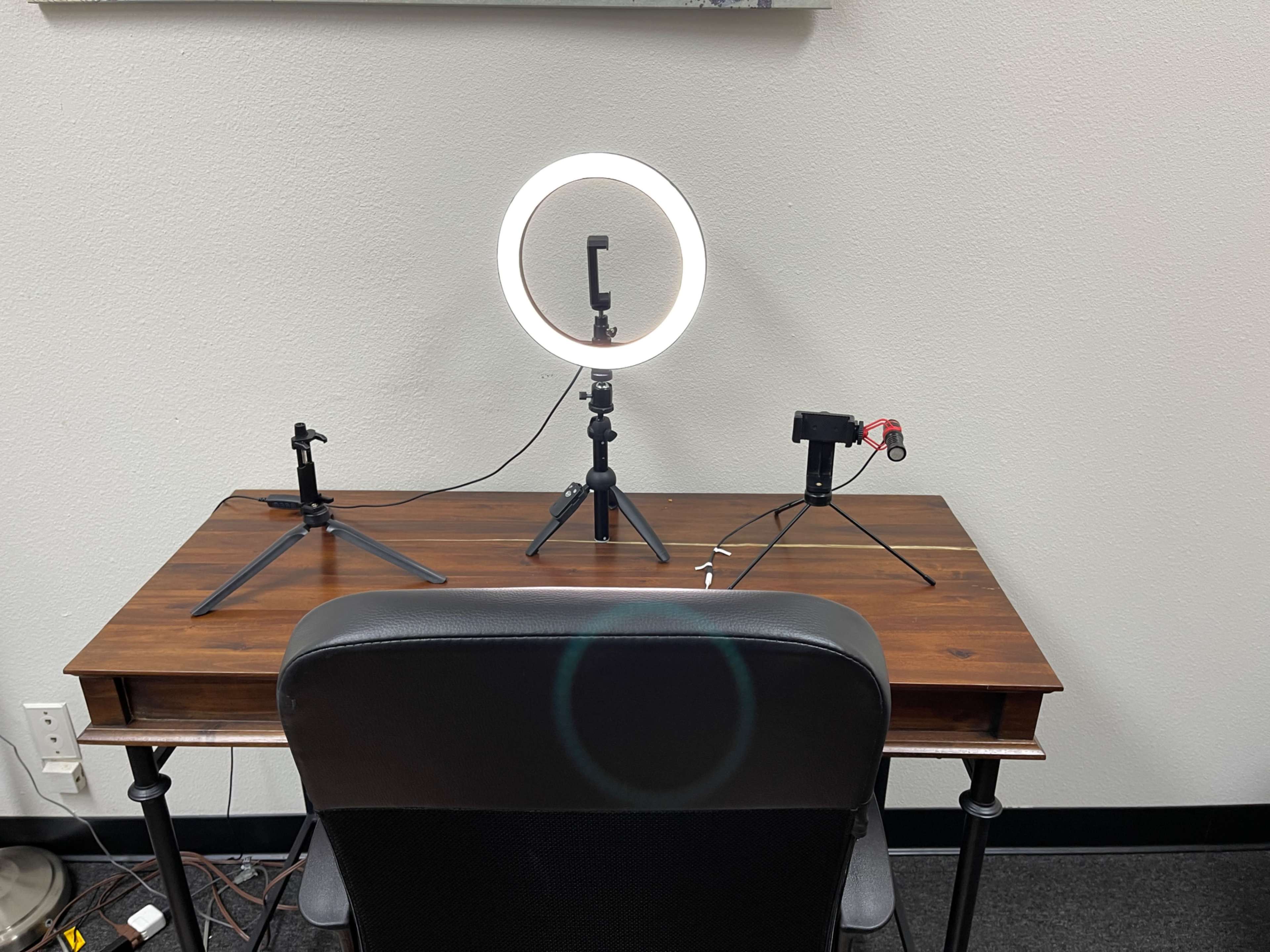 A wooden desk is set up with a ring light and two tripod stands for equipment, with a black chair positioned in front.
