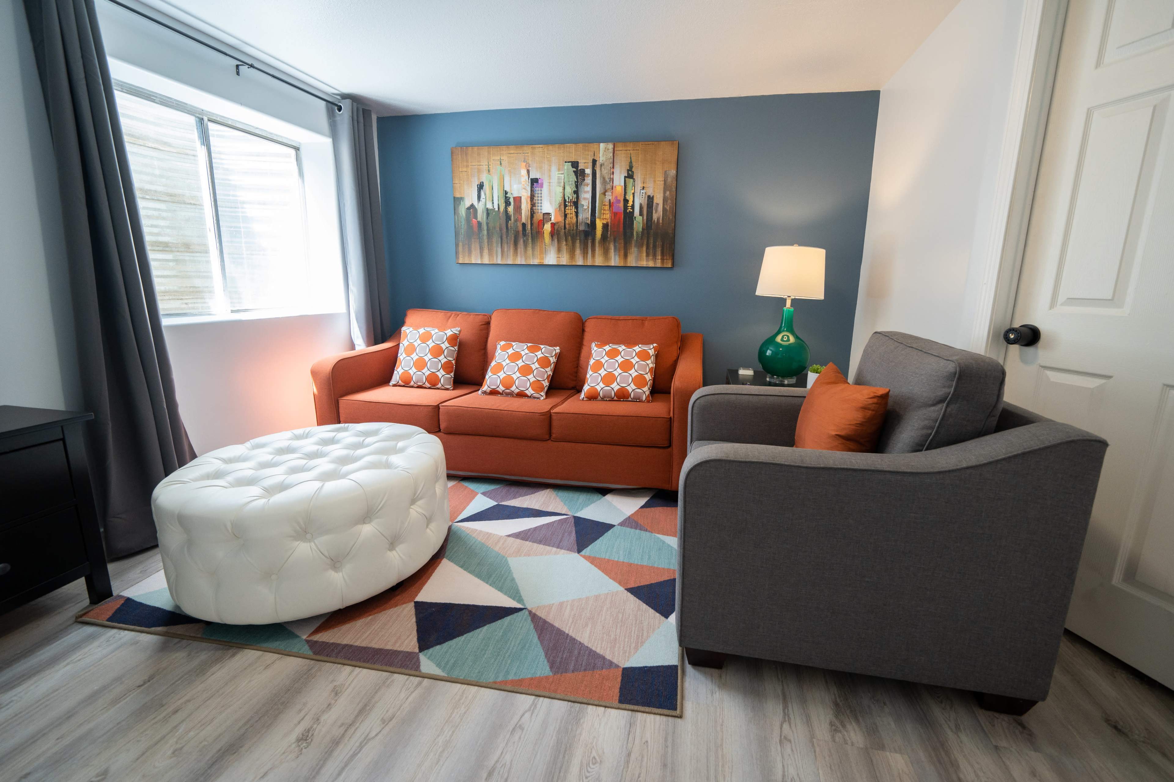 The living room features an orange sofa with decorative pillows, a gray armchair, a round white ottoman, and a geometric-patterned rug, all arranged around a window with natural light.