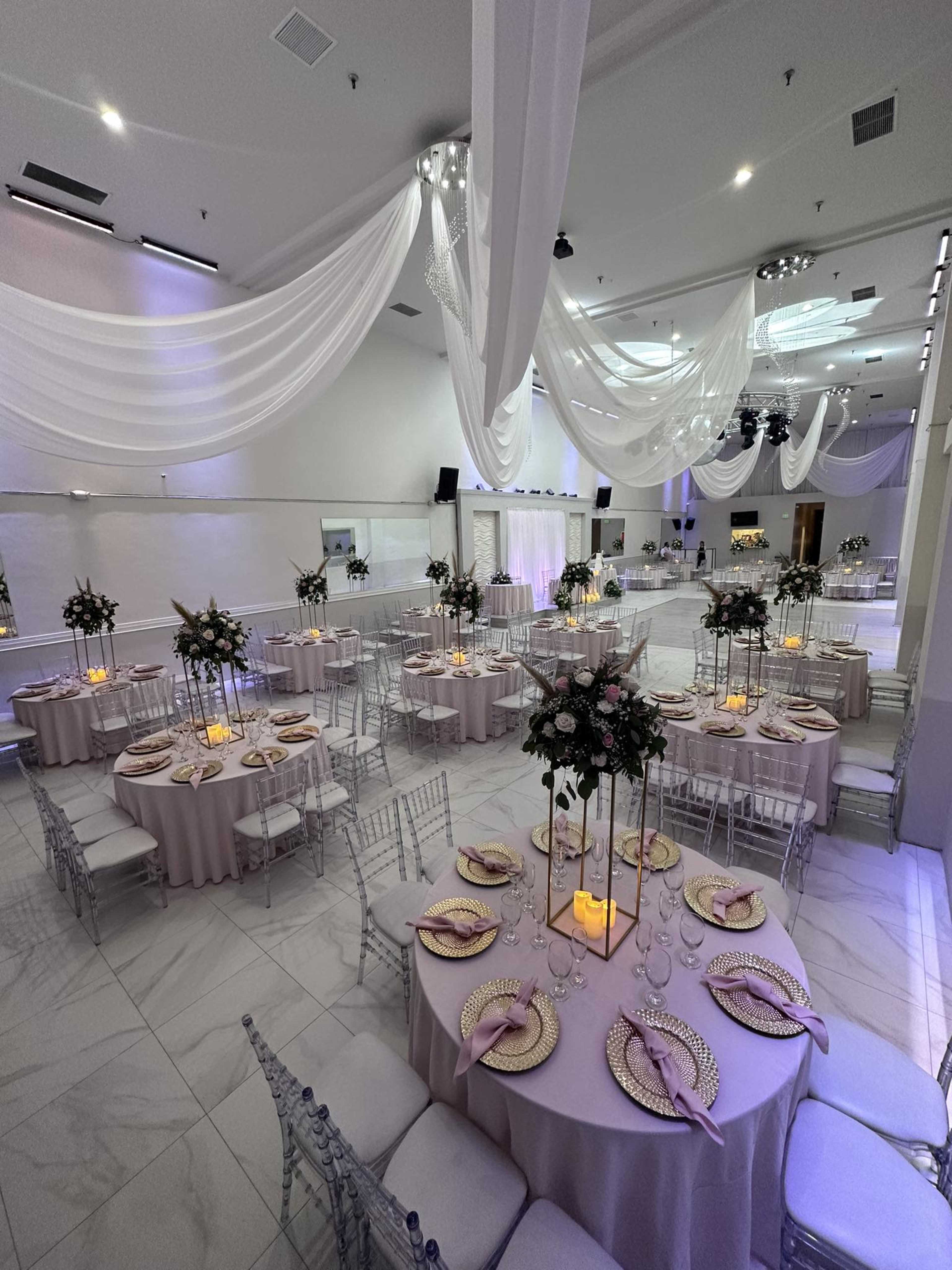 The image shows a banquet hall decorated for an event, featuring round tables with pink tablecloths, elegant centerpieces, and draped white fabric across the ceiling.
