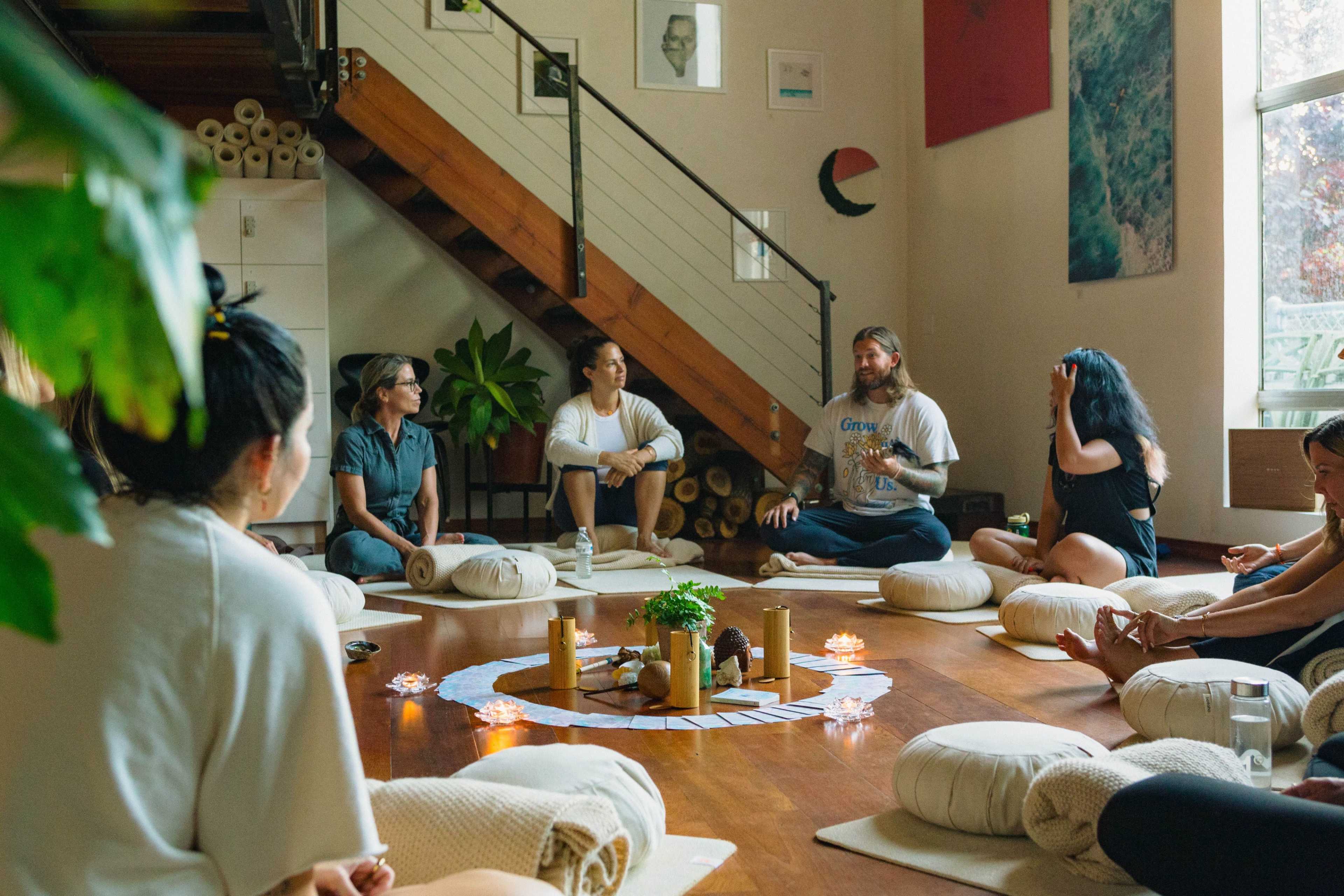 A group of individuals sits in a circle on the floor of a well-lit room, engaging in a discussion surrounded by decorative pillows and candles.