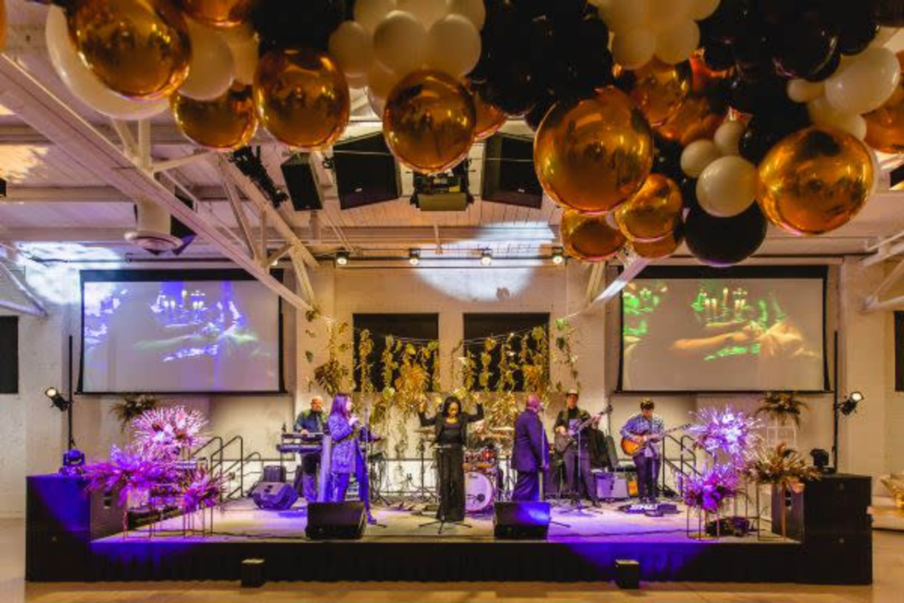 A band performs on stage in a decorated venue with large screens and black and gold balloon arrangements overhead.