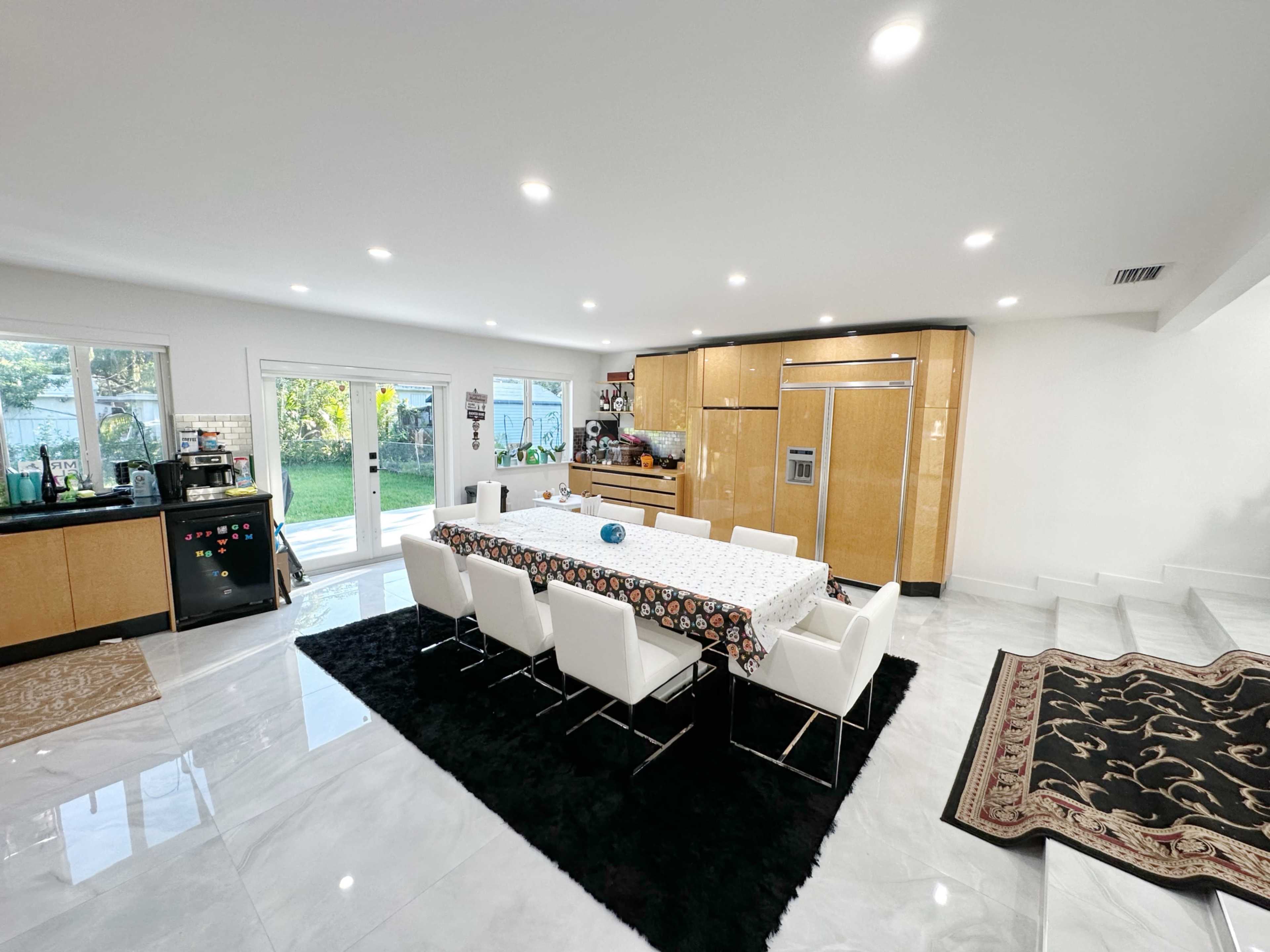 A modern kitchen and dining area features a large table with white chairs, sleek cabinetry, and sliding glass doors leading to a backyard.