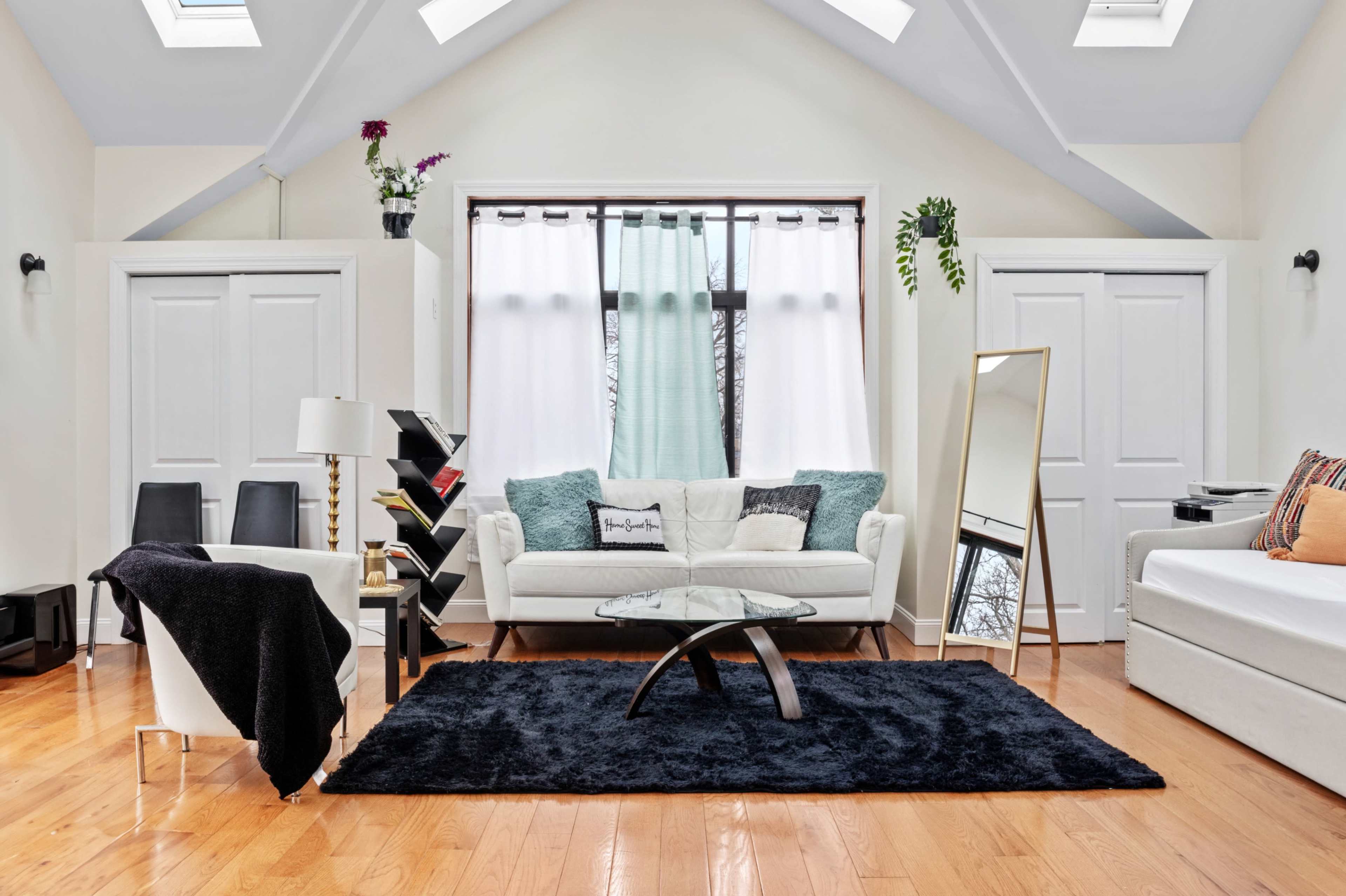 The room features a light-colored sofa, a glass coffee table, and a large mirror, all positioned on a dark rug over polished wooden floors, with doors on either side and windows letting in natural light.