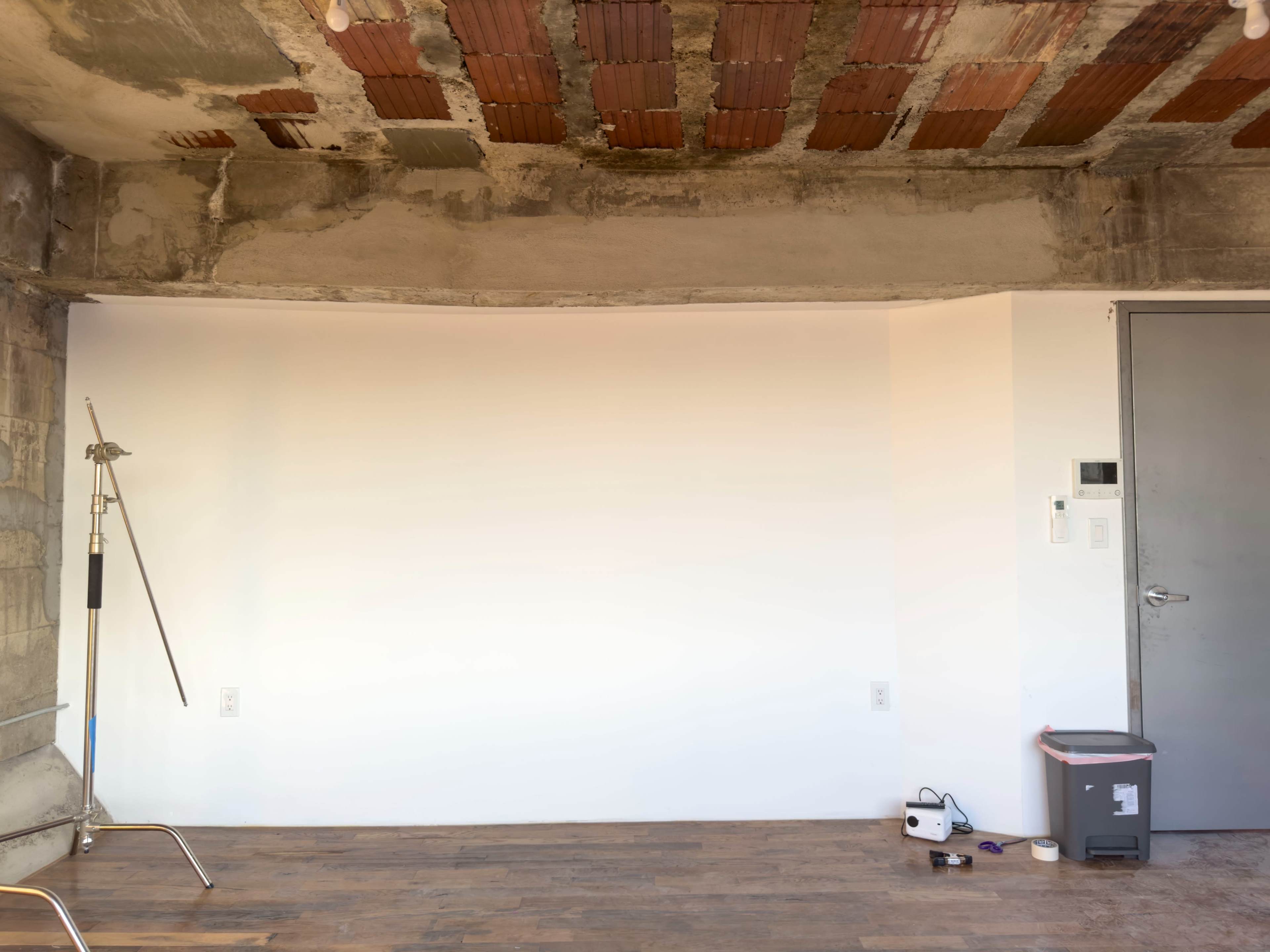 An unfinished interior space with exposed brick and concrete walls, a light wooden floor, and minimal furnishings including a trash can and a small device on the floor.