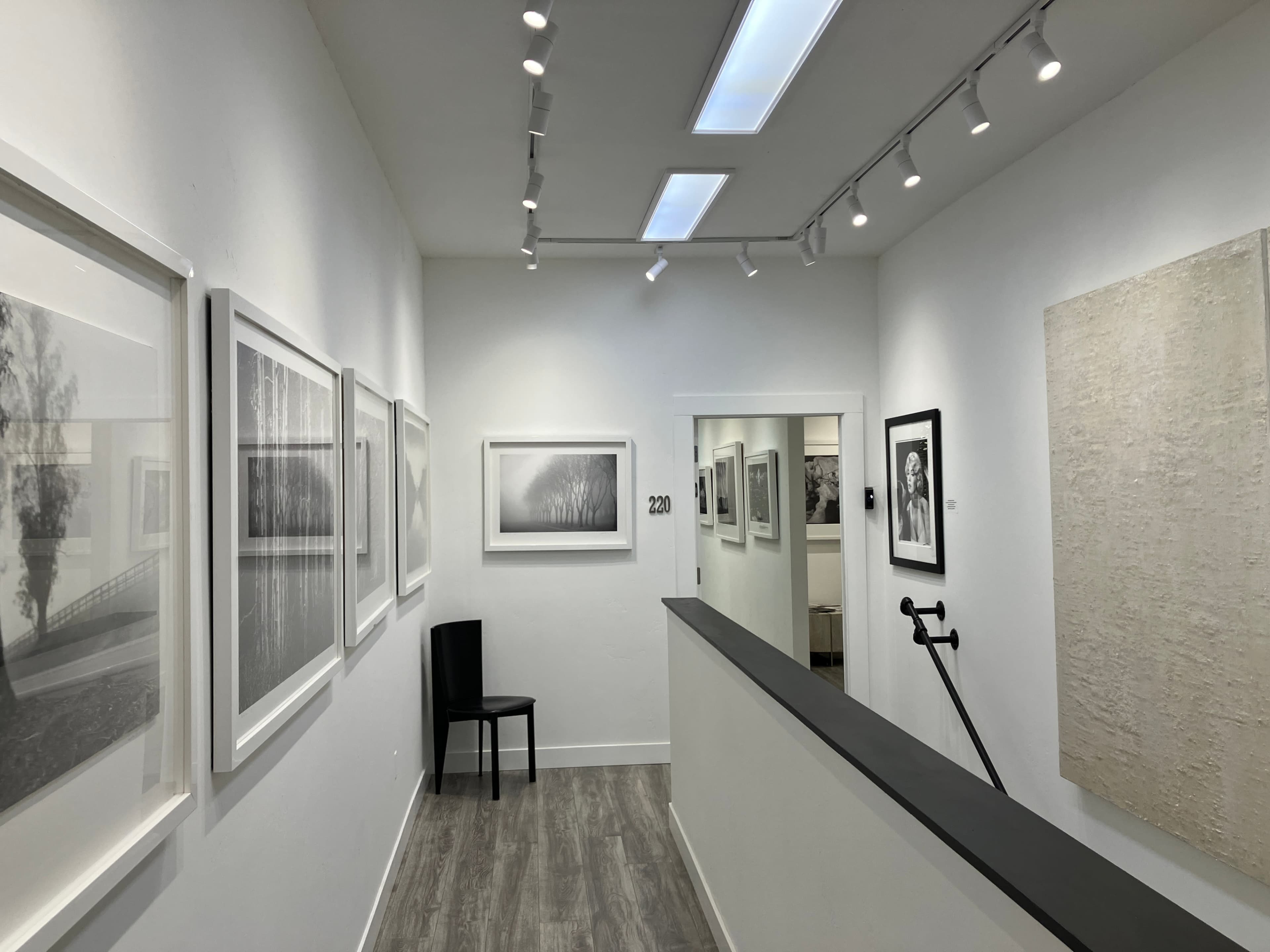 A gallery hallway features framed black-and-white photographs lining the walls, with a single chair positioned near the entrance.