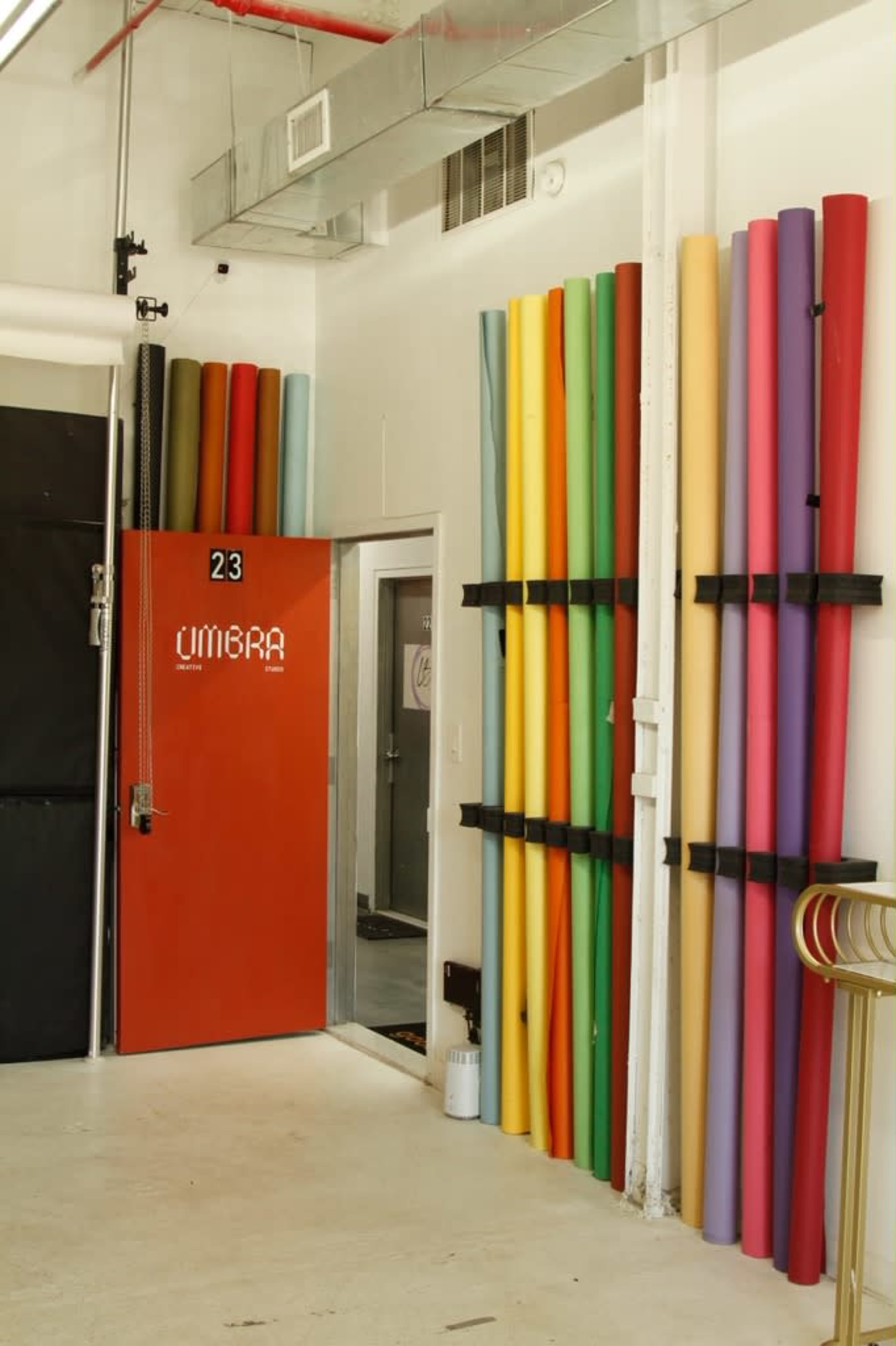 The image shows a room with colorful rolls of paper stacked vertically against the wall, and a door marked "23" painted in bright orange.