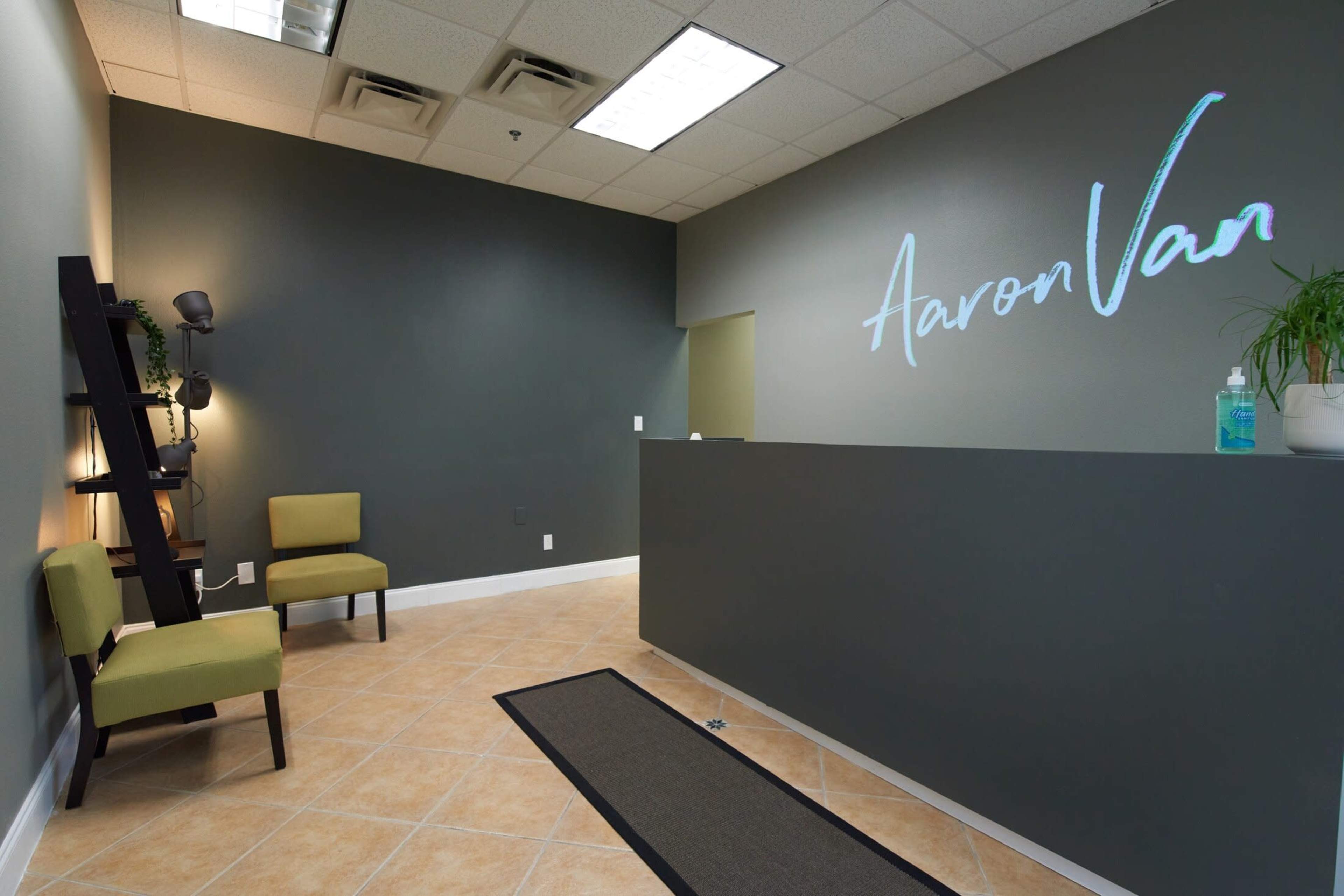 The image shows a modern reception area with a desk, two green chairs, and a decorative ladder shelf against a gray wall featuring the name "Aaron Van."
