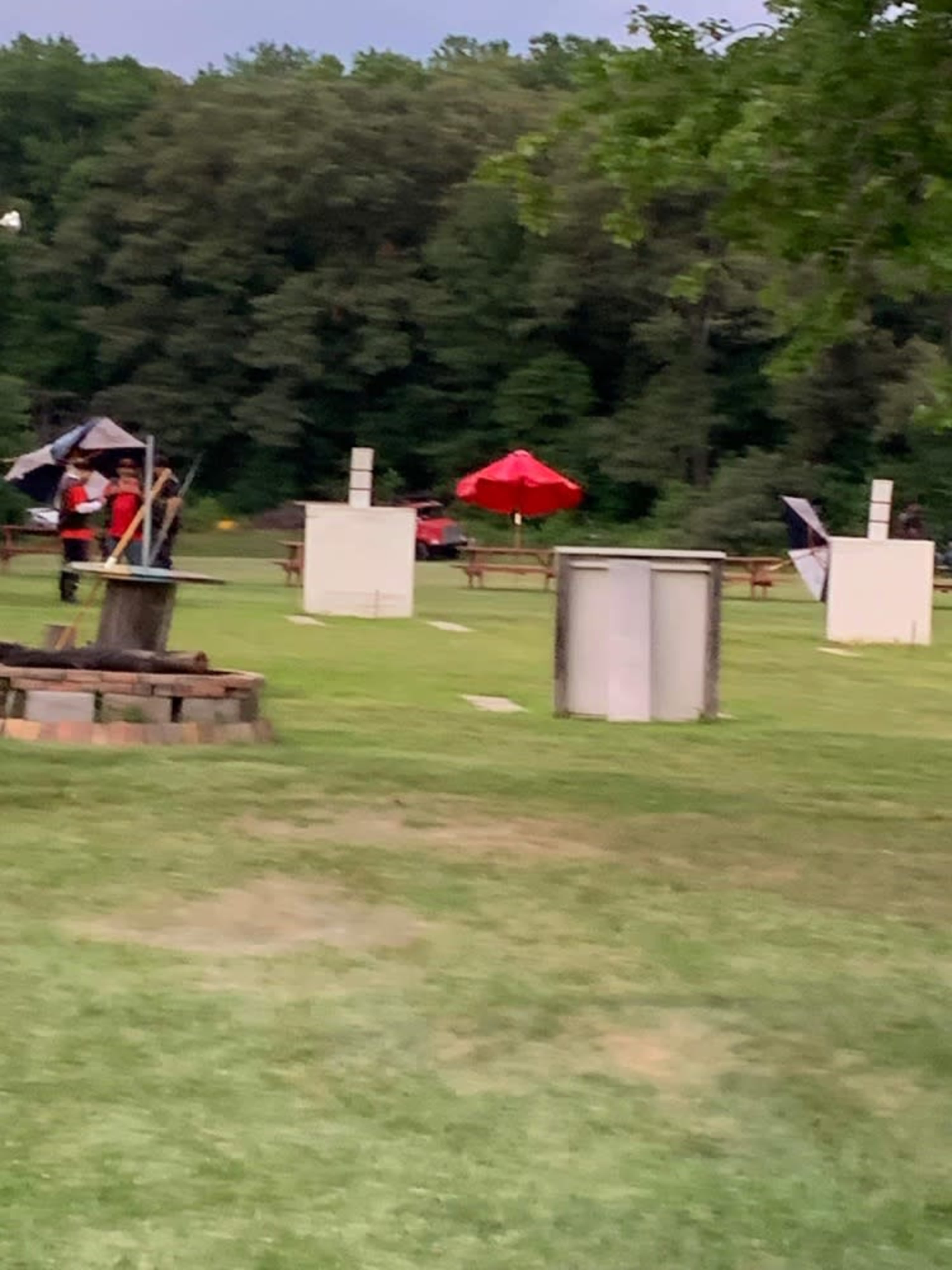 A grassy area with picnic tables, white targets, and people with umbrellas in the background.