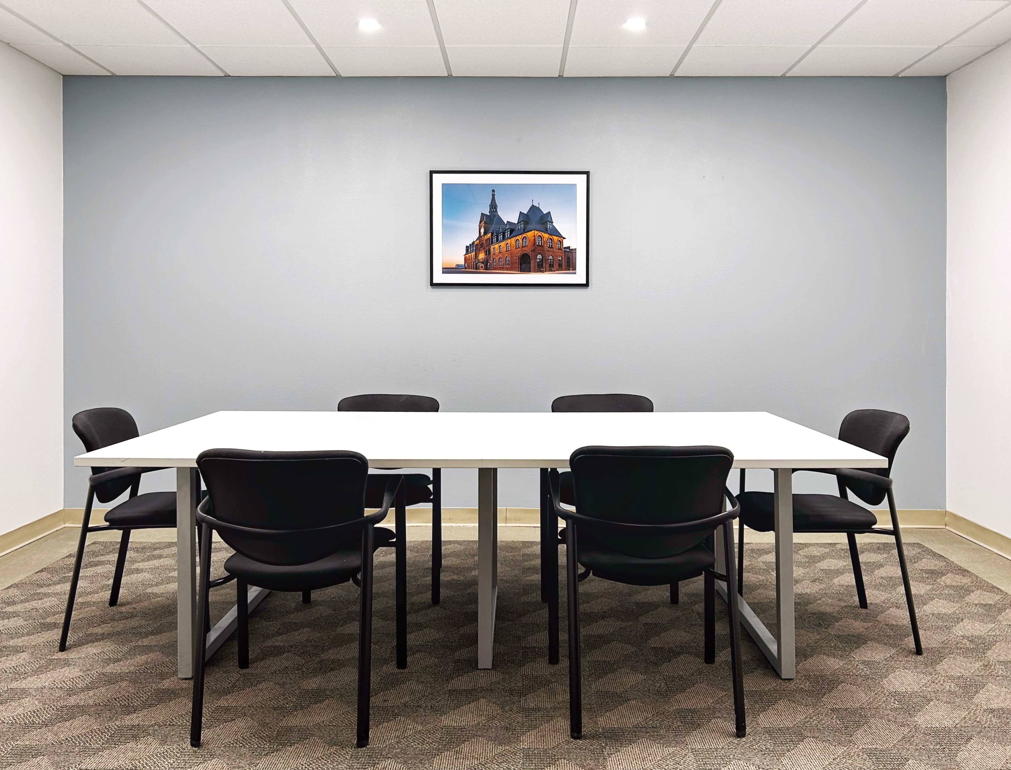 A modern conference room features a large white table surrounded by six black chairs, with a framed picture of a building on the wall.