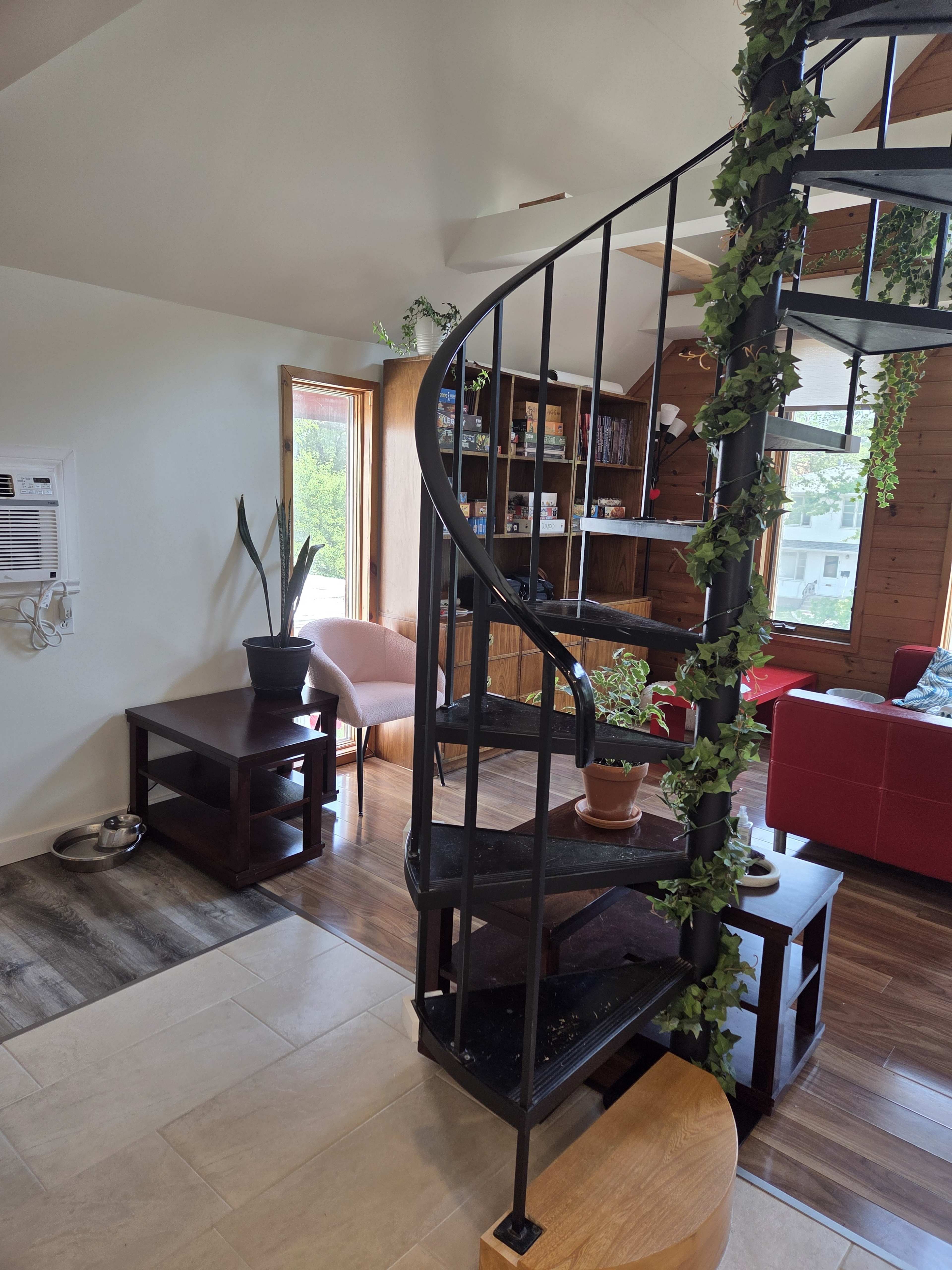 The image shows a modern interior with a spiral staircase, wooden shelves filled with books, and a cozy seating area with a red couch.