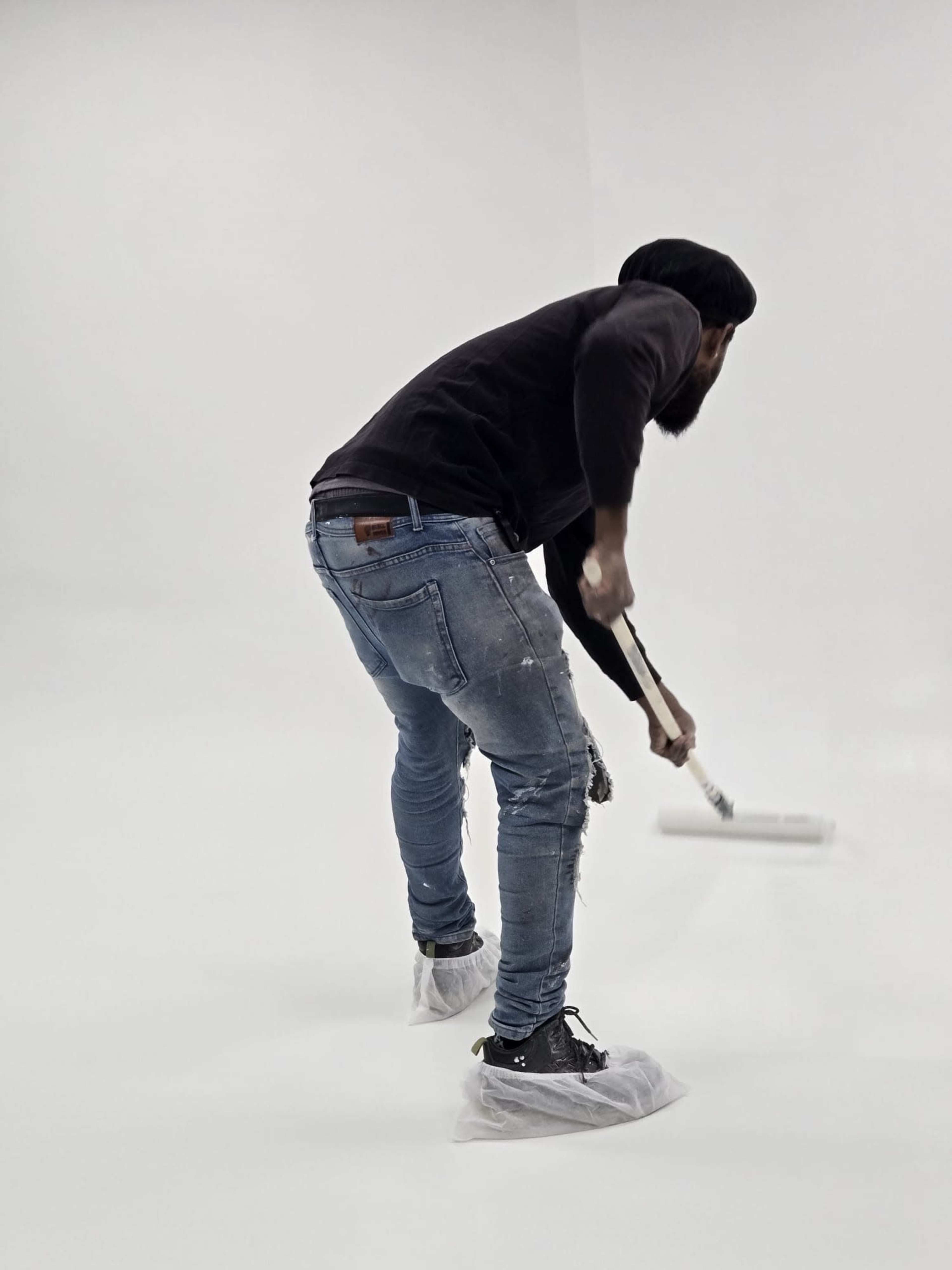 A person in jeans and a black top is painting a white wall with a roller while wearing protective shoe covers.