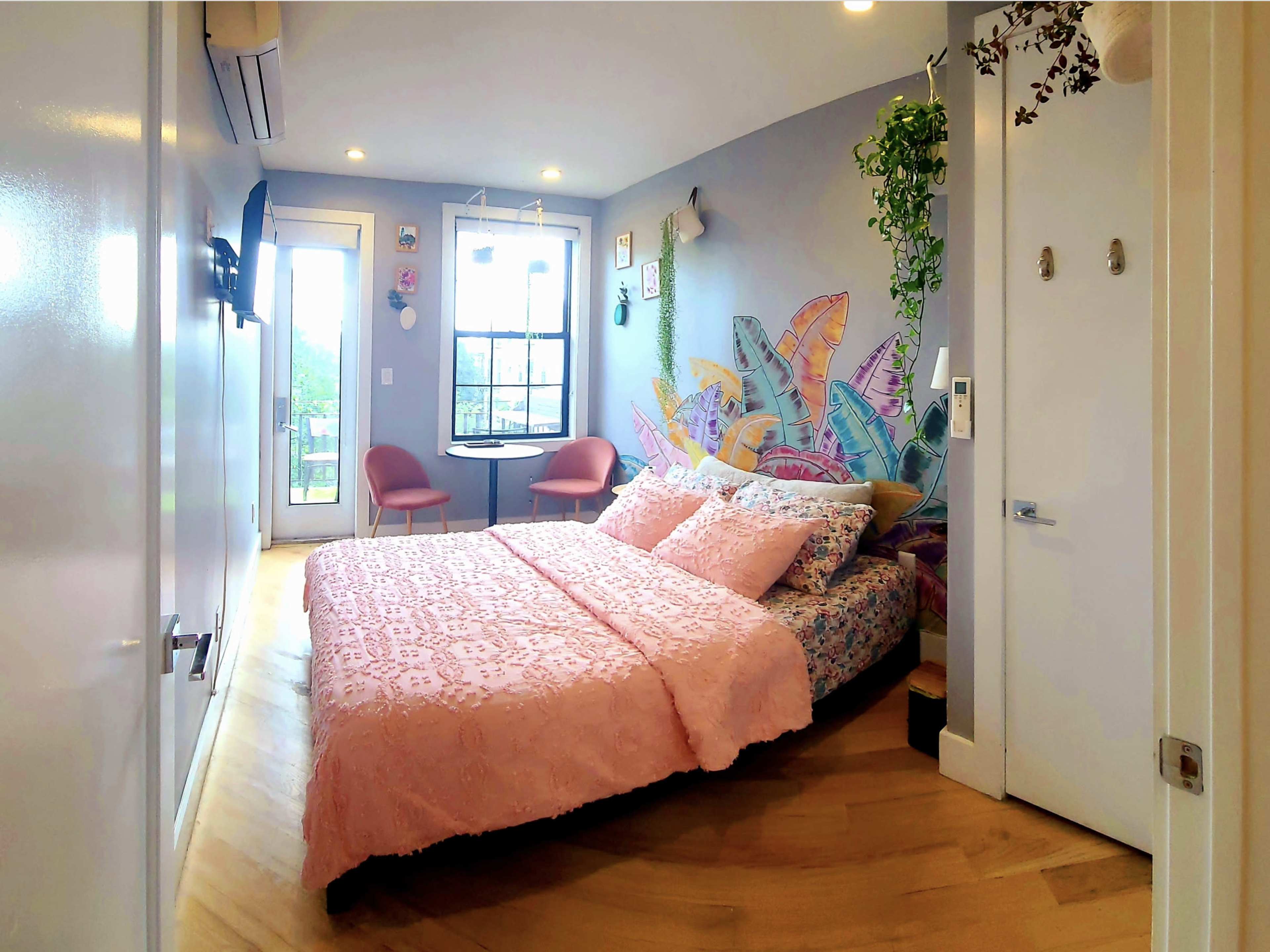 A cozy bedroom featuring a bed with a pink quilt, a small table with chairs, and colorful wall art adorned with plants.