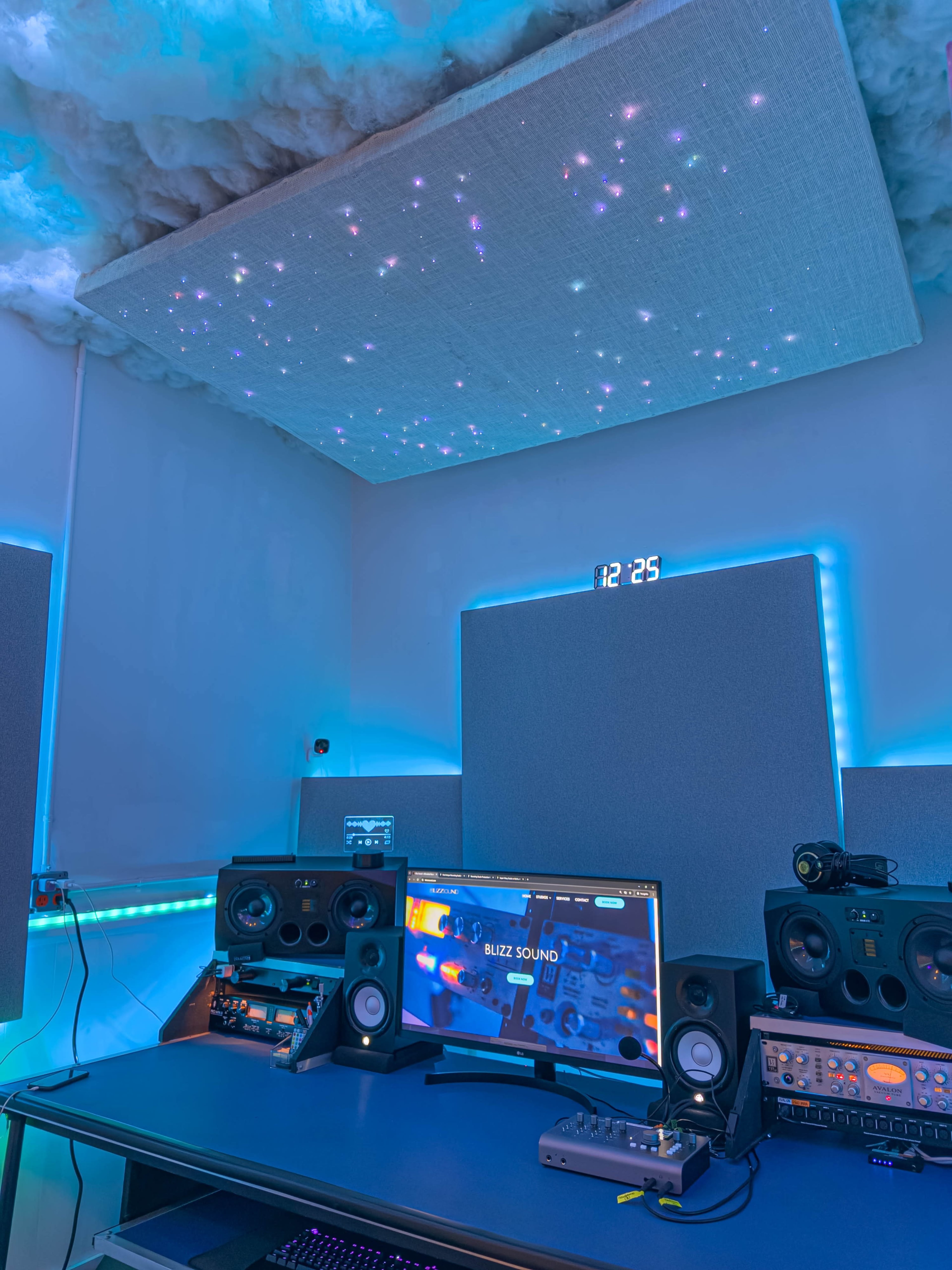 The image shows a modern music studio setup with sound equipment, a computer monitor displaying a music software interface, and a cloud-like acoustic ceiling with small lights, all illuminated by blue LED accents.