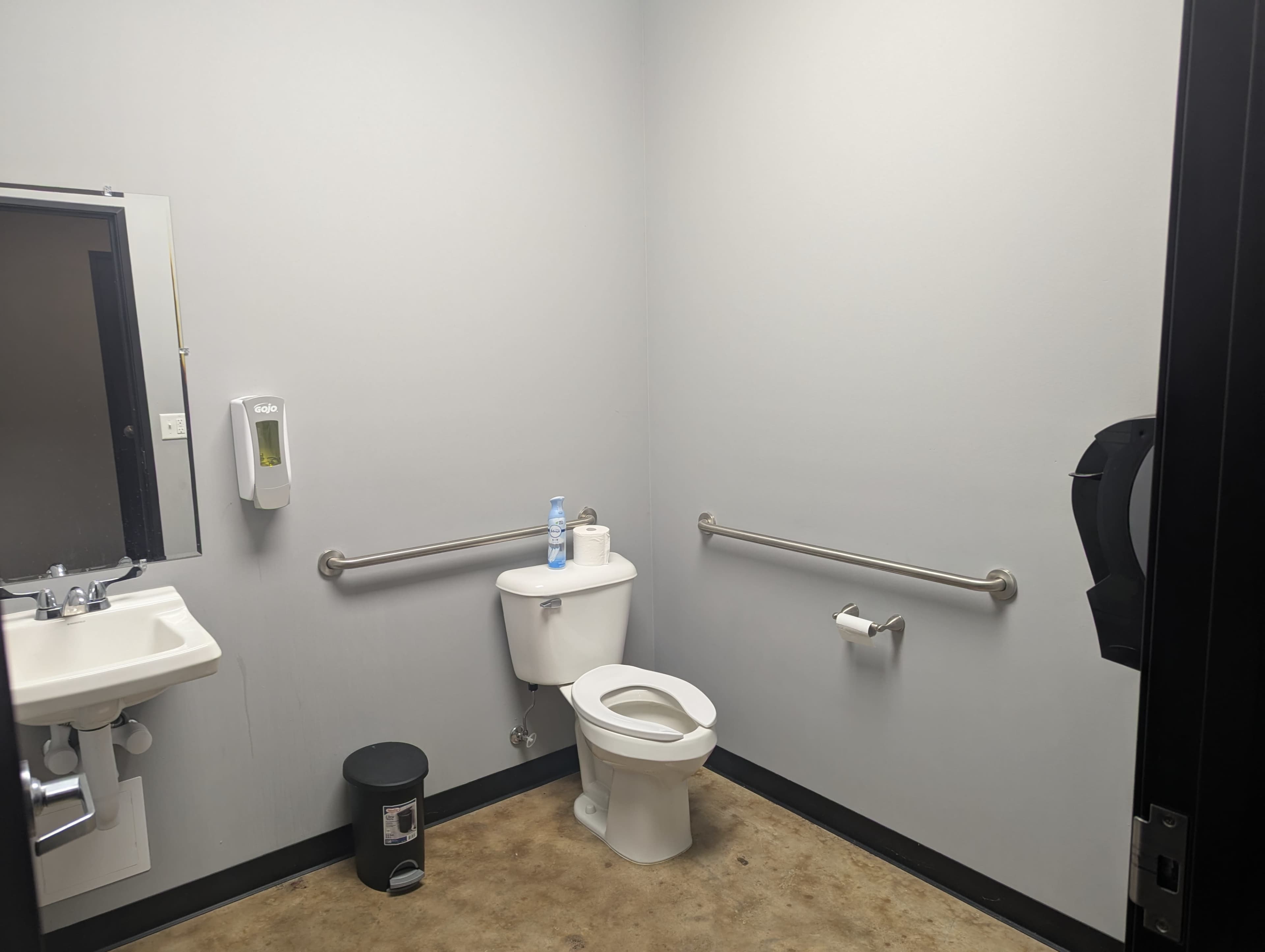 The image shows a small restroom with a toilet, sink, hand sanitizer dispenser, and a trash can.