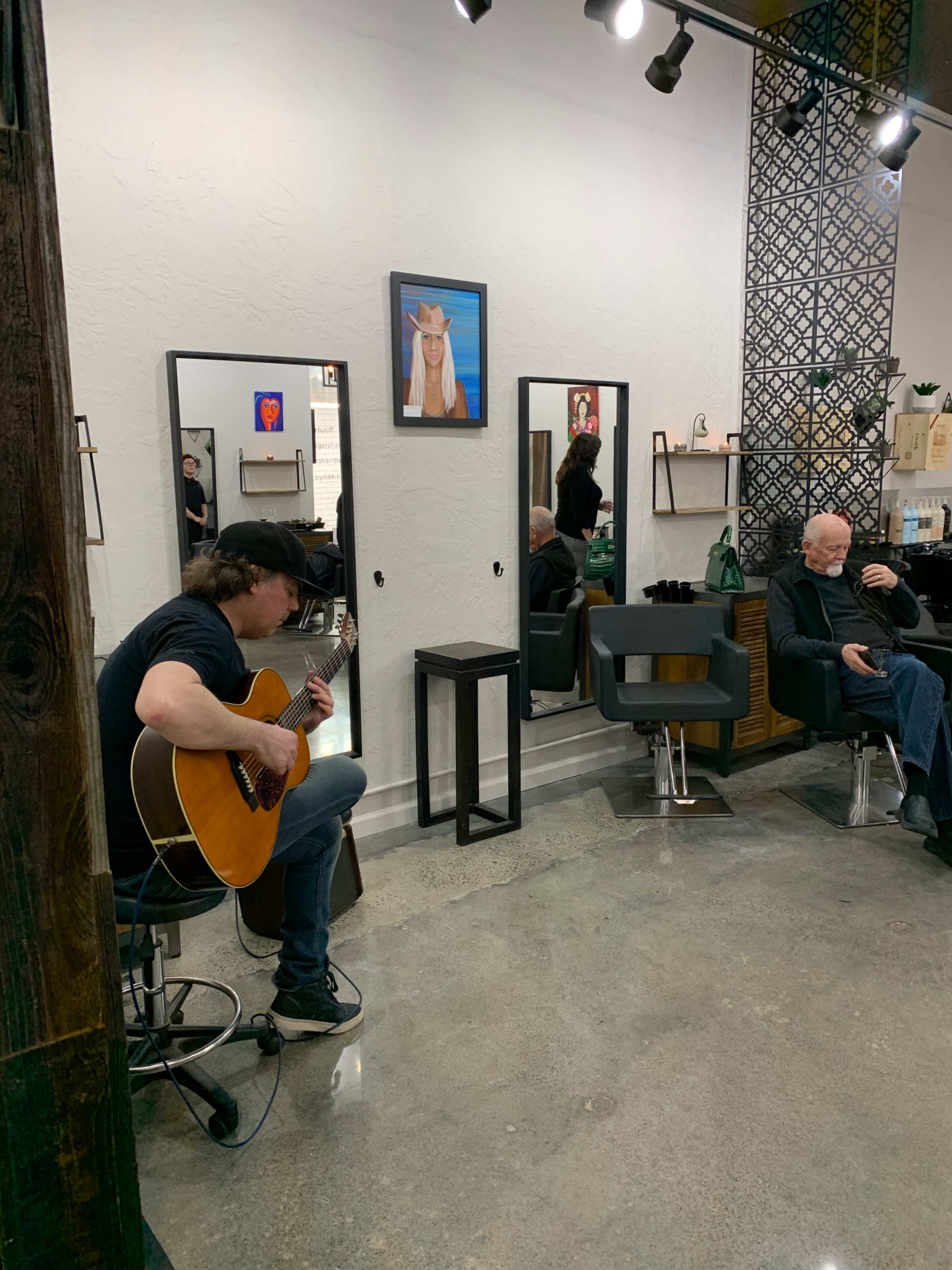 A musician plays guitar in a hair salon while a client sits in a chair getting a haircut.