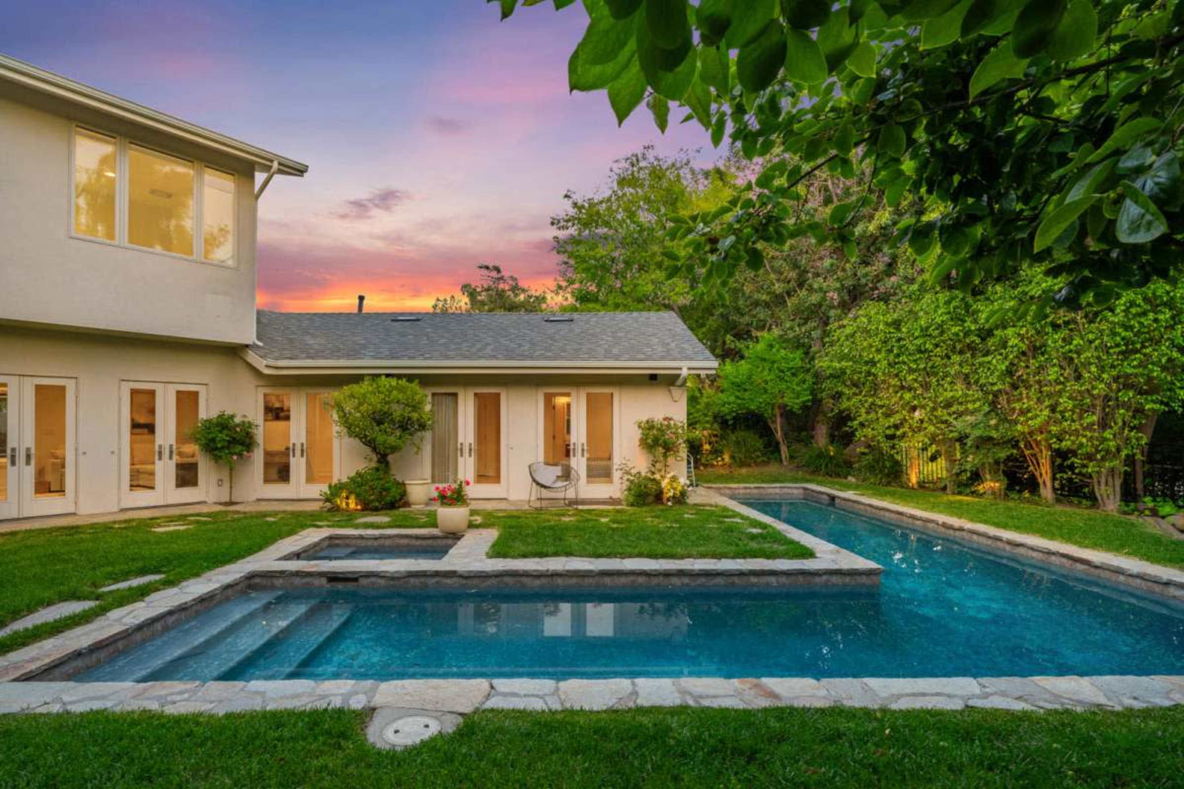 The image shows a backyard with a swimming pool, landscaped greenery, and a home with multiple doors, set against a colorful sunset sky.