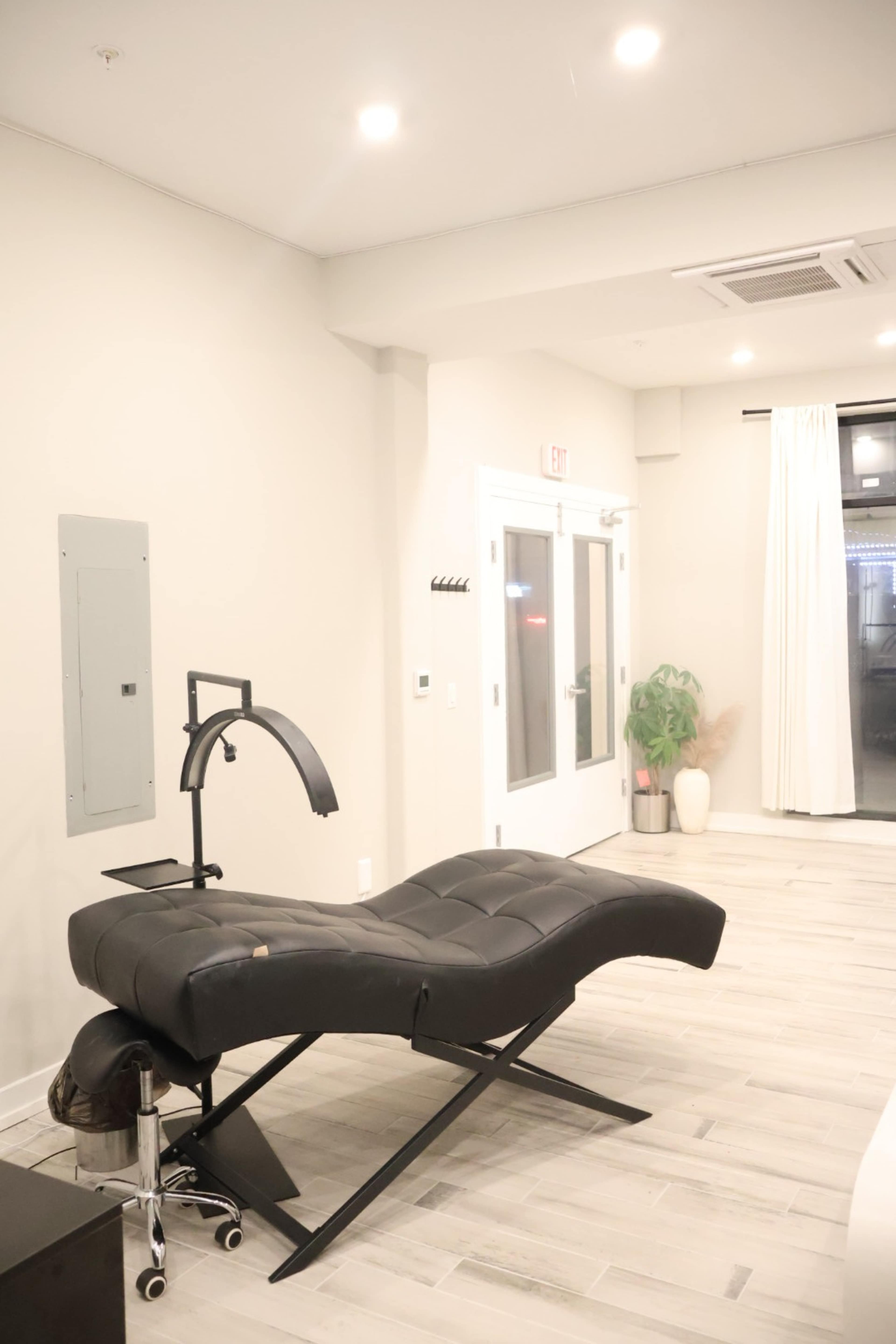 The image shows a modern treatment room featuring a black adjustable reclining chair, a small rolling cart, and large windows with sheer curtains.