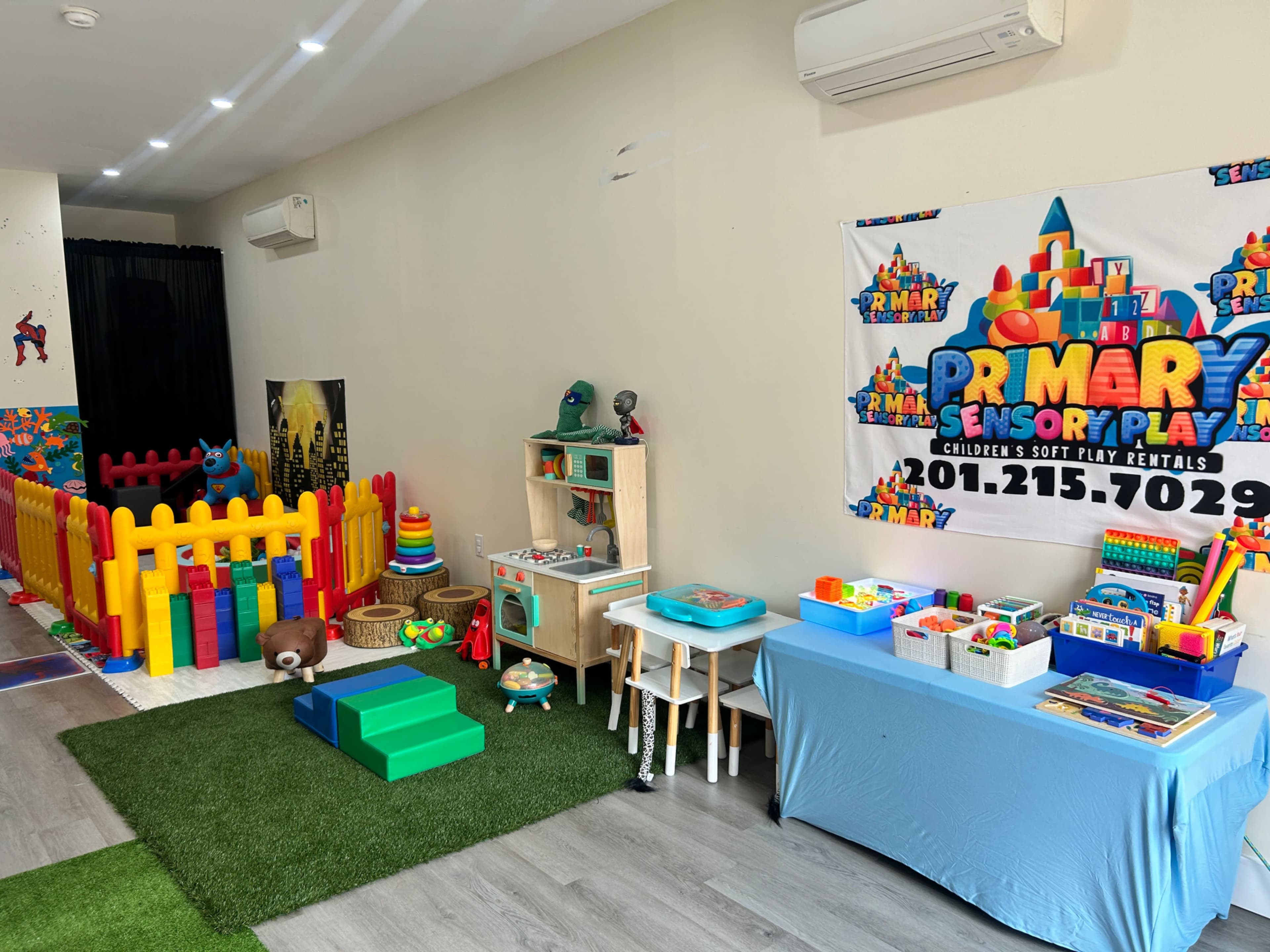 The image shows a colorful children's play area featuring a fenced play zone, various toys, and an activity table, along with a promotional banner for a soft play rental service.