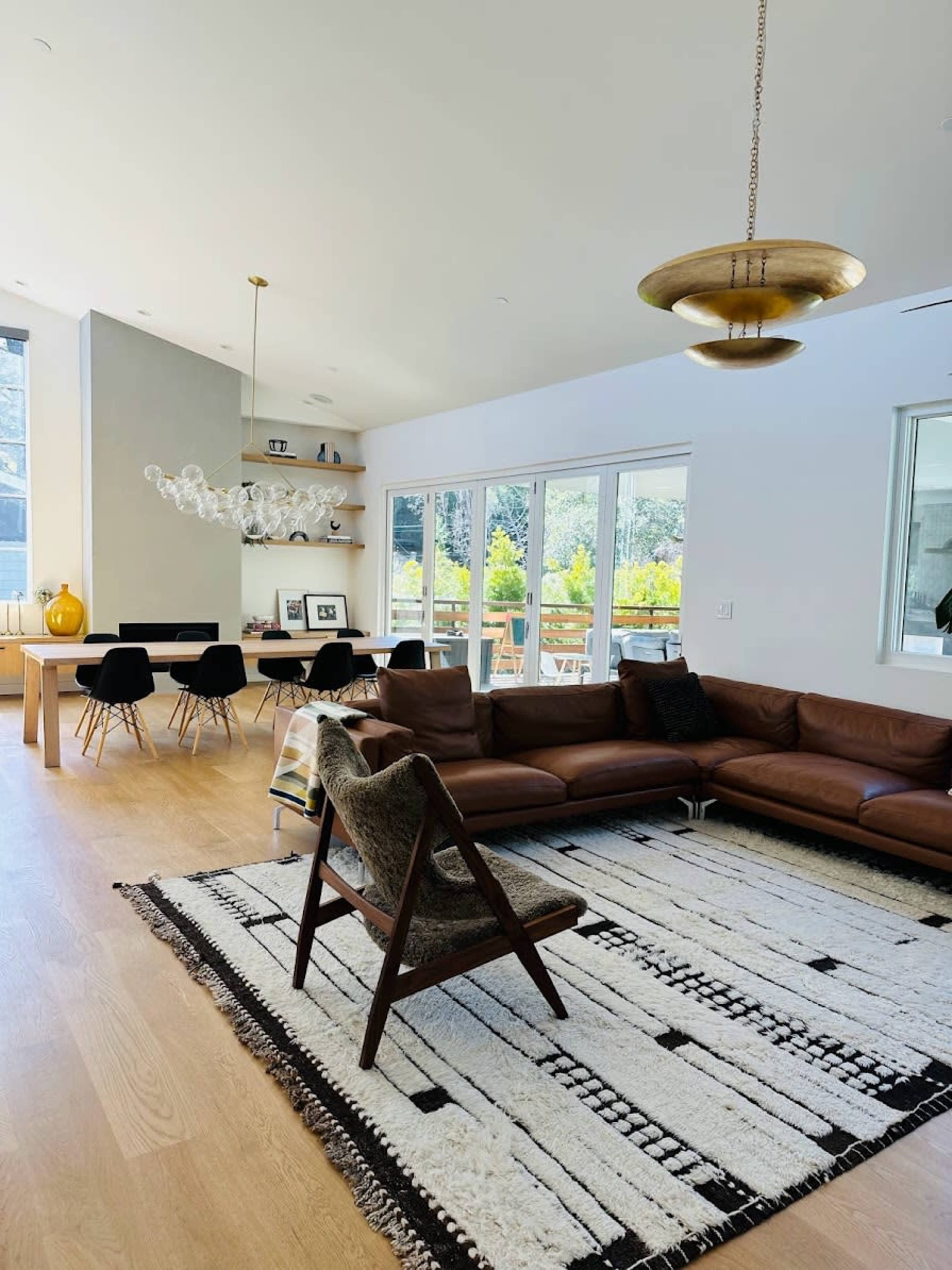 A modern living space featuring a brown leather sectional sofa, a patterned rug, a dining table with black chairs, and large windows that allow natural light.