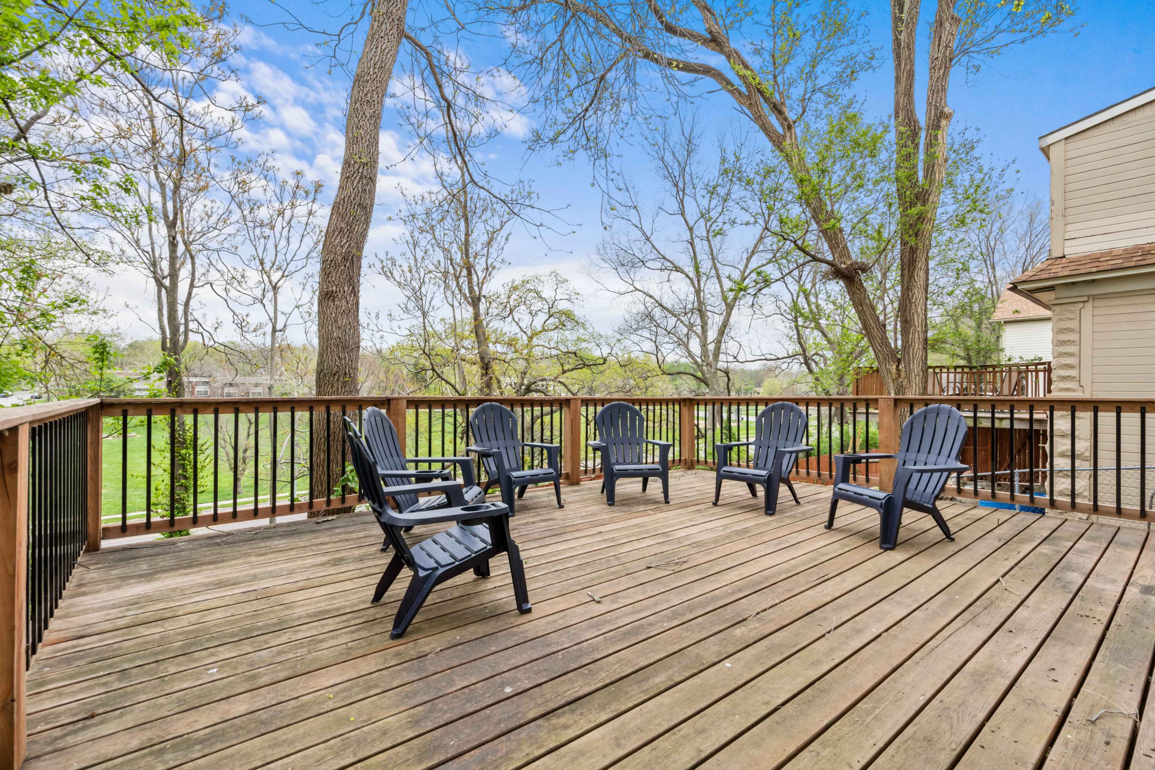 A wooden deck with five adirondack chairs overlooks a grassy area and trees.