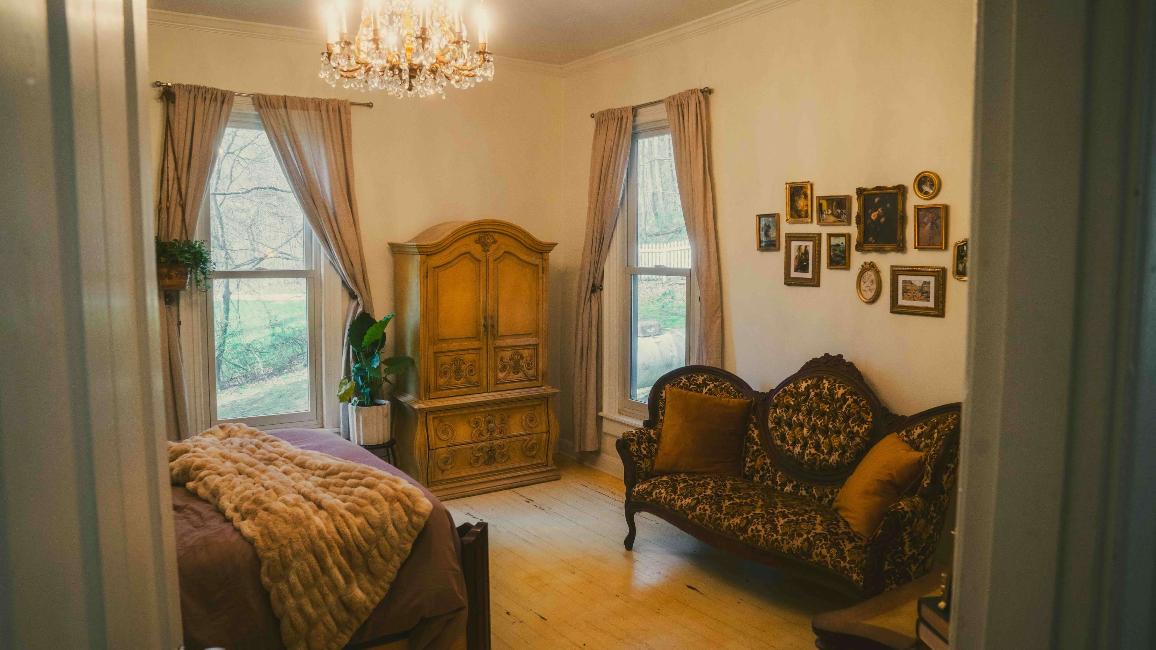 The image shows a softly lit bedroom featuring a vintage wooden bed, a decorative sofa, a large wardrobe, and various framed pictures on the wall.