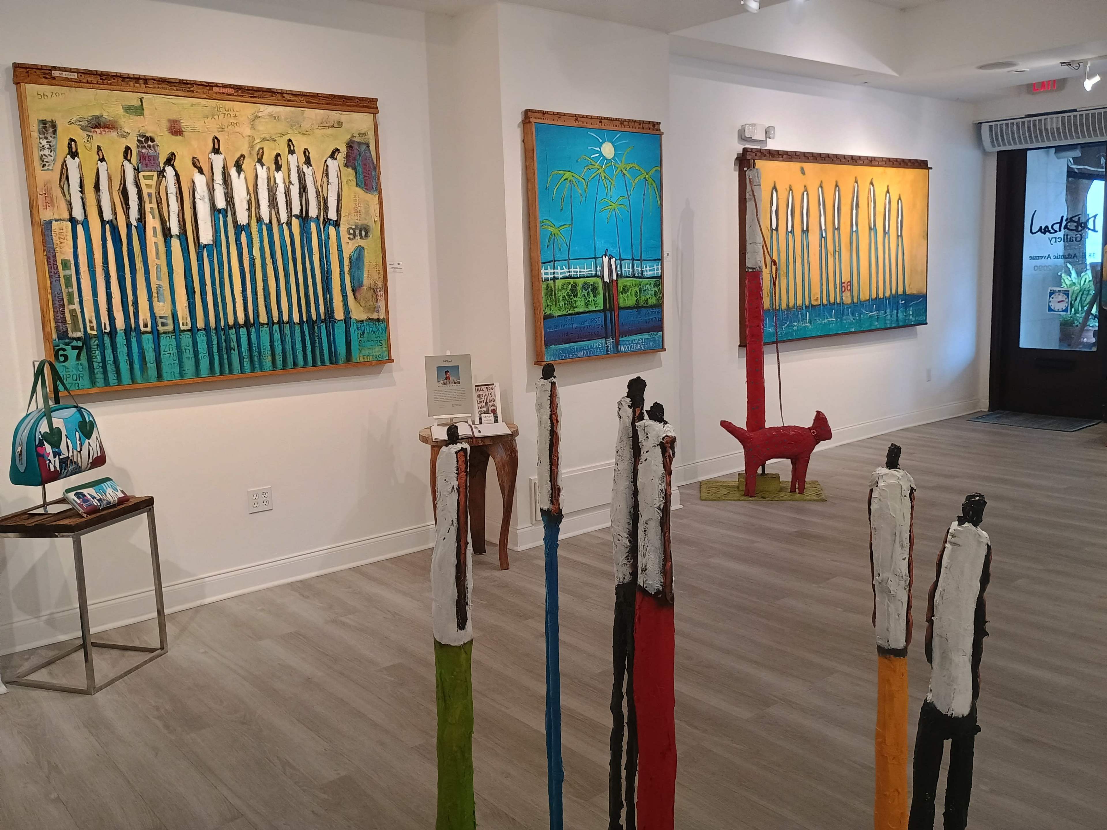 The image features an art gallery interior with several colorful paintings on the walls and sculptures resembling tall, painted figures positioned in the foreground.