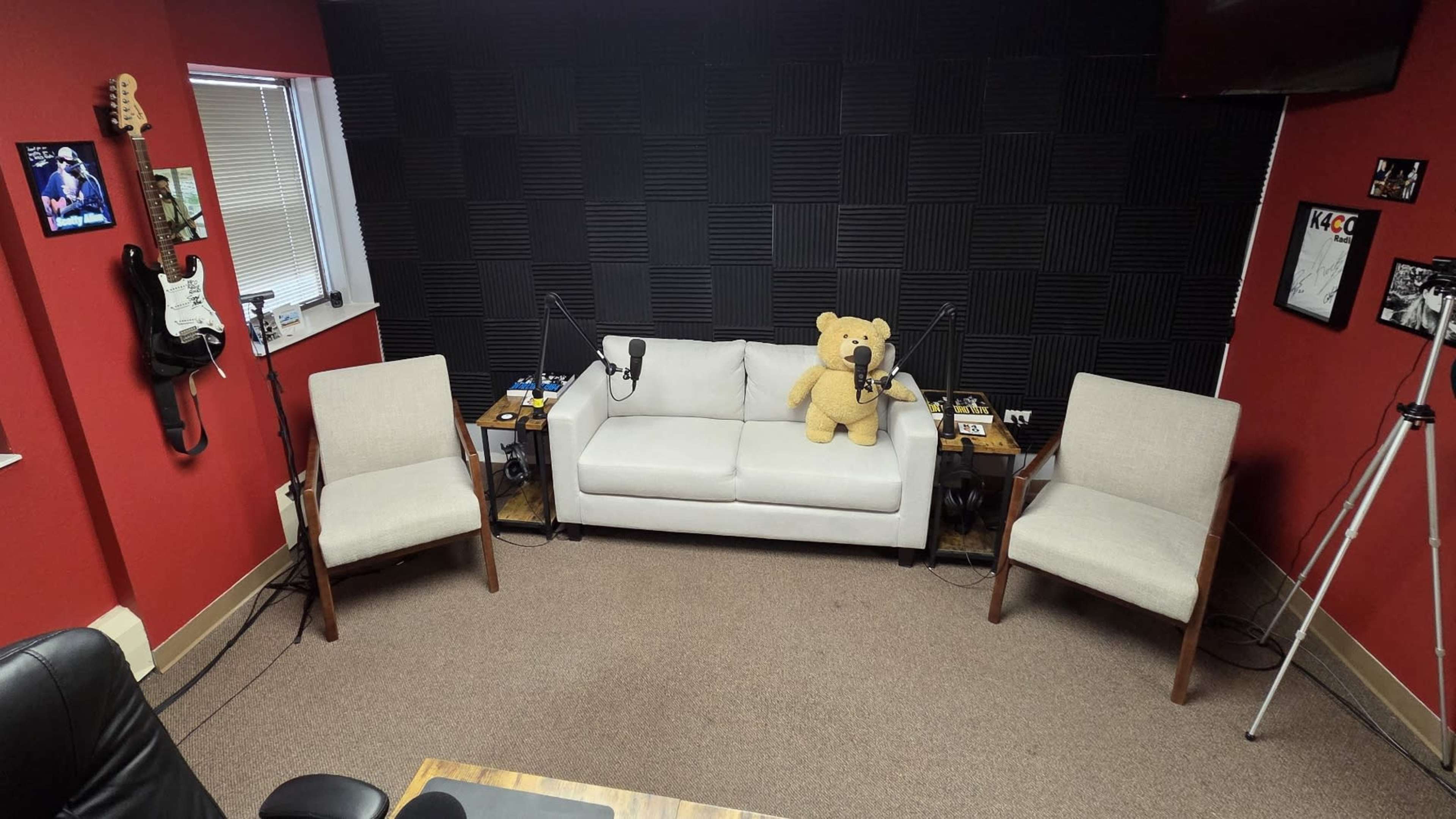 The image shows a small recording studio with a white sofa, two beige chairs, and microphones set up on stands, all against a backdrop of black soundproofing panels on the walls.